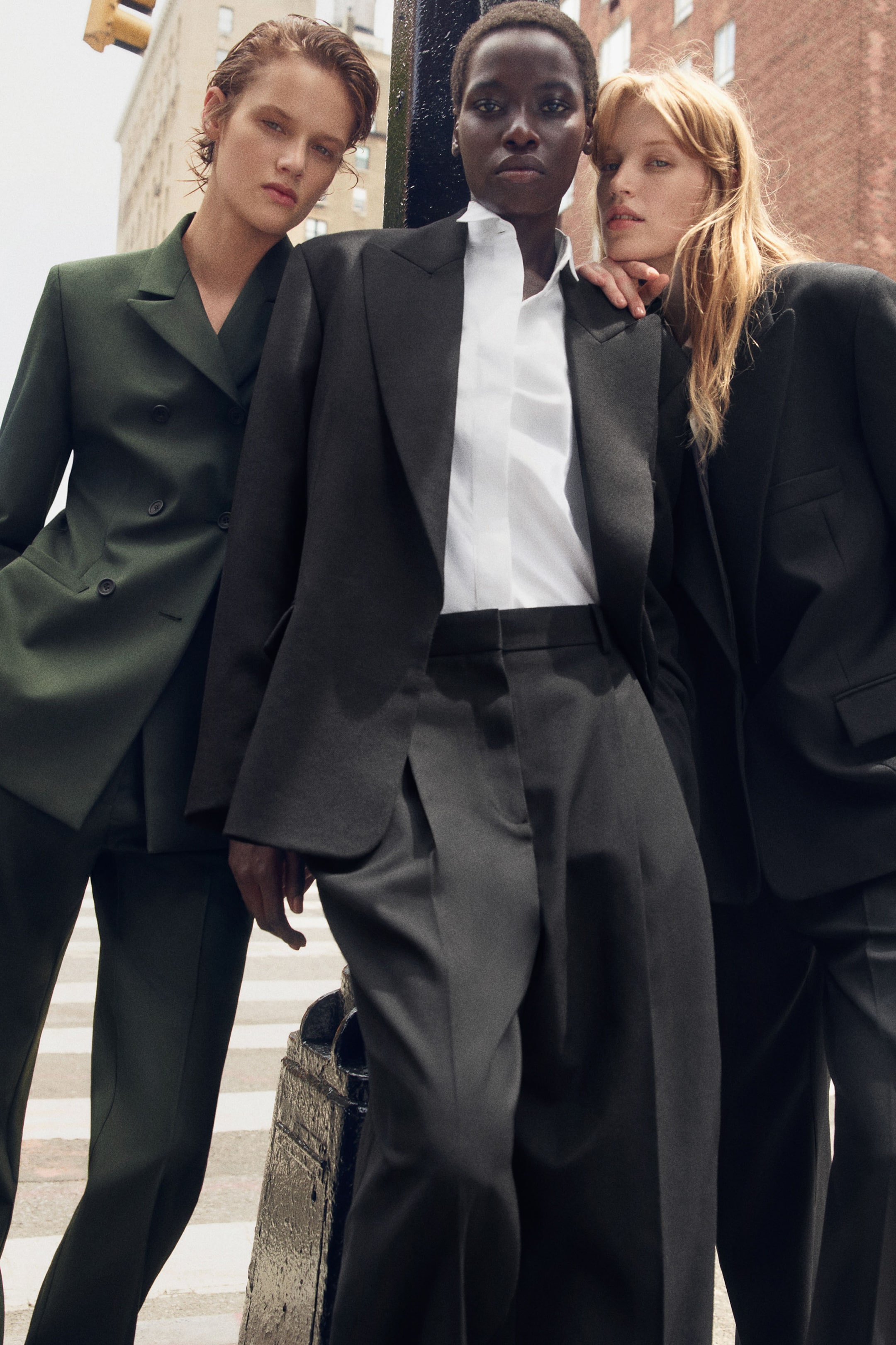 Agrandir l'image: Un mannequin porte une veste en laine noire cintrée et tailleur, avec épaules structurées et revers à pointes, portée sur une chemise de smoking en coton blanche à col cassé. La tenue est complétée par un pantalon évasé noir assorti, taille haute, en twill tailleur, avec plis sur le devant.