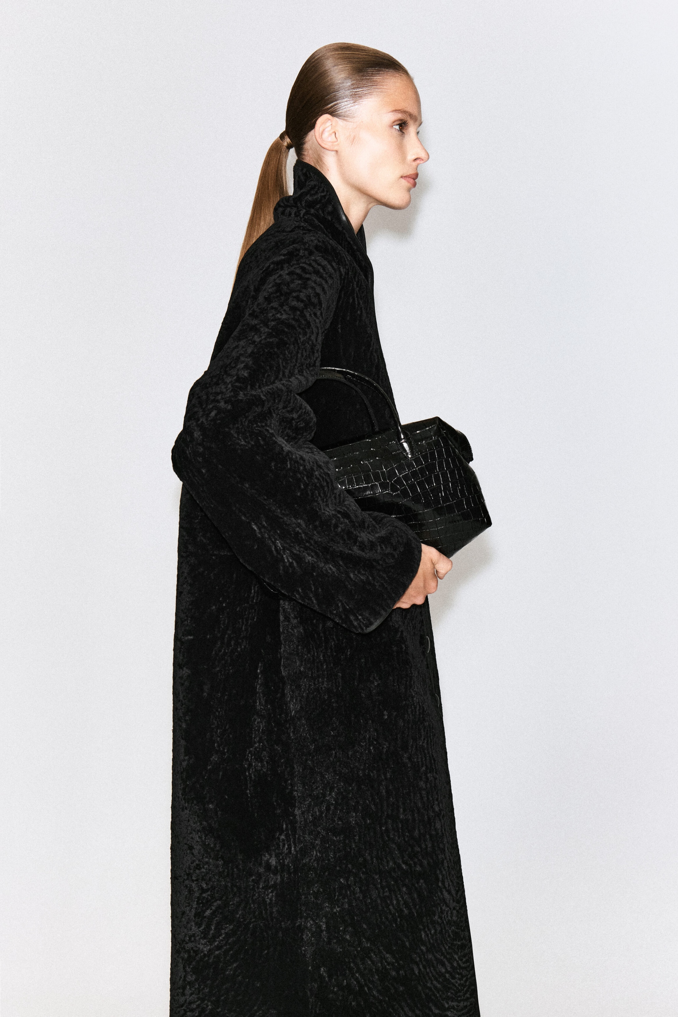 A black croc-debossed leather bowling bag with a sculptural shape and top handles is being carried by the model, who holds it close to their body. The model is wearing a textured black coat that coordinates with the bag. A black croc-debossed leather bowling bag with a sculptural shape and top handles is being carried by the model, who holds it close to their body. The model is wearing a textured black coat that coordinates with the bag.