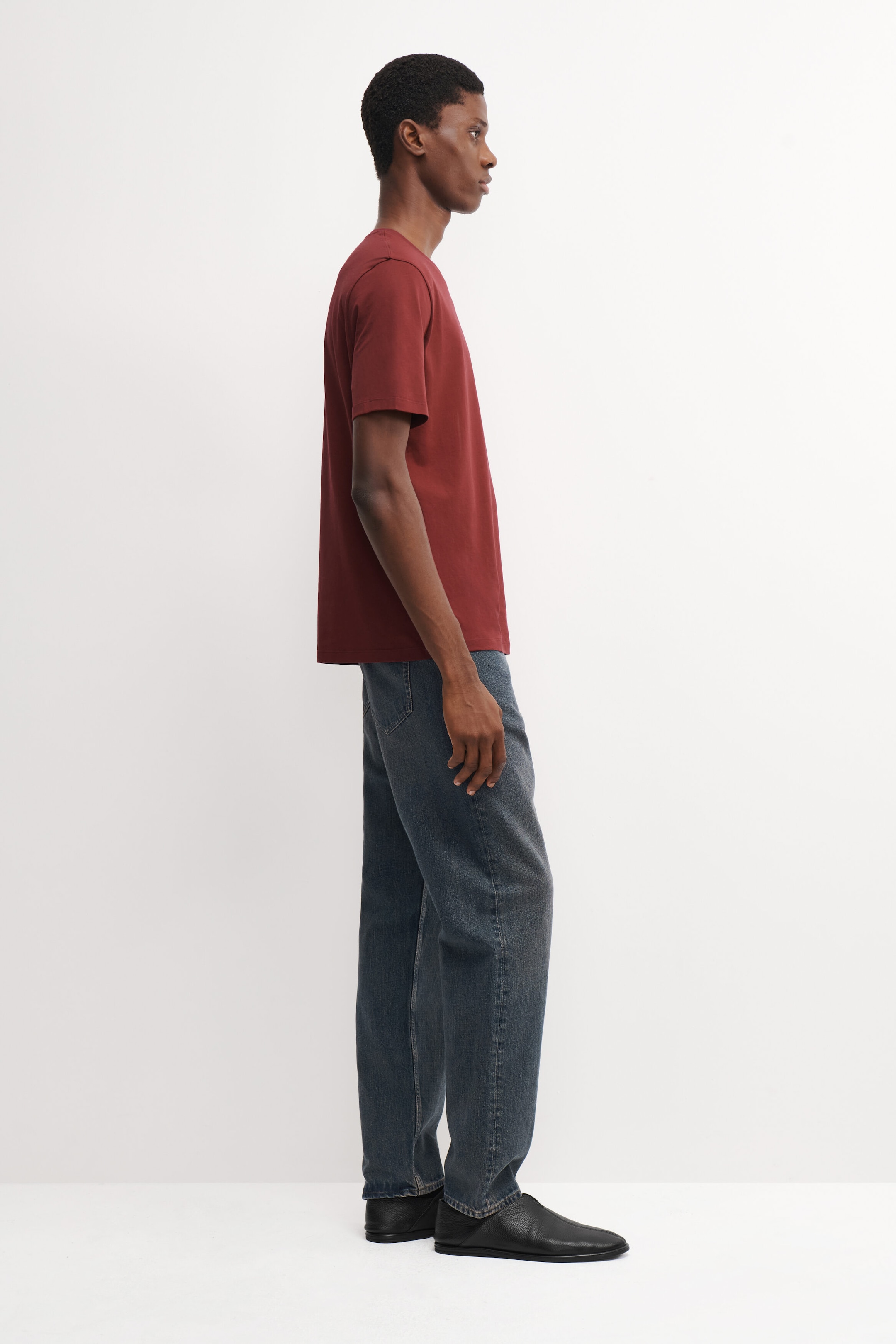 A dark red cotton crew-neck T-shirt is worn by the model, who is standing in side profile. The short-sleeved T-shirt is styled with dark gray denim jeans and black slip-on shoes. A dark red cotton crew-neck T-shirt is worn by the model, who is standing in side profile. The short-sleeved T-shirt is styled with dark gray denim jeans and black slip-on shoes.