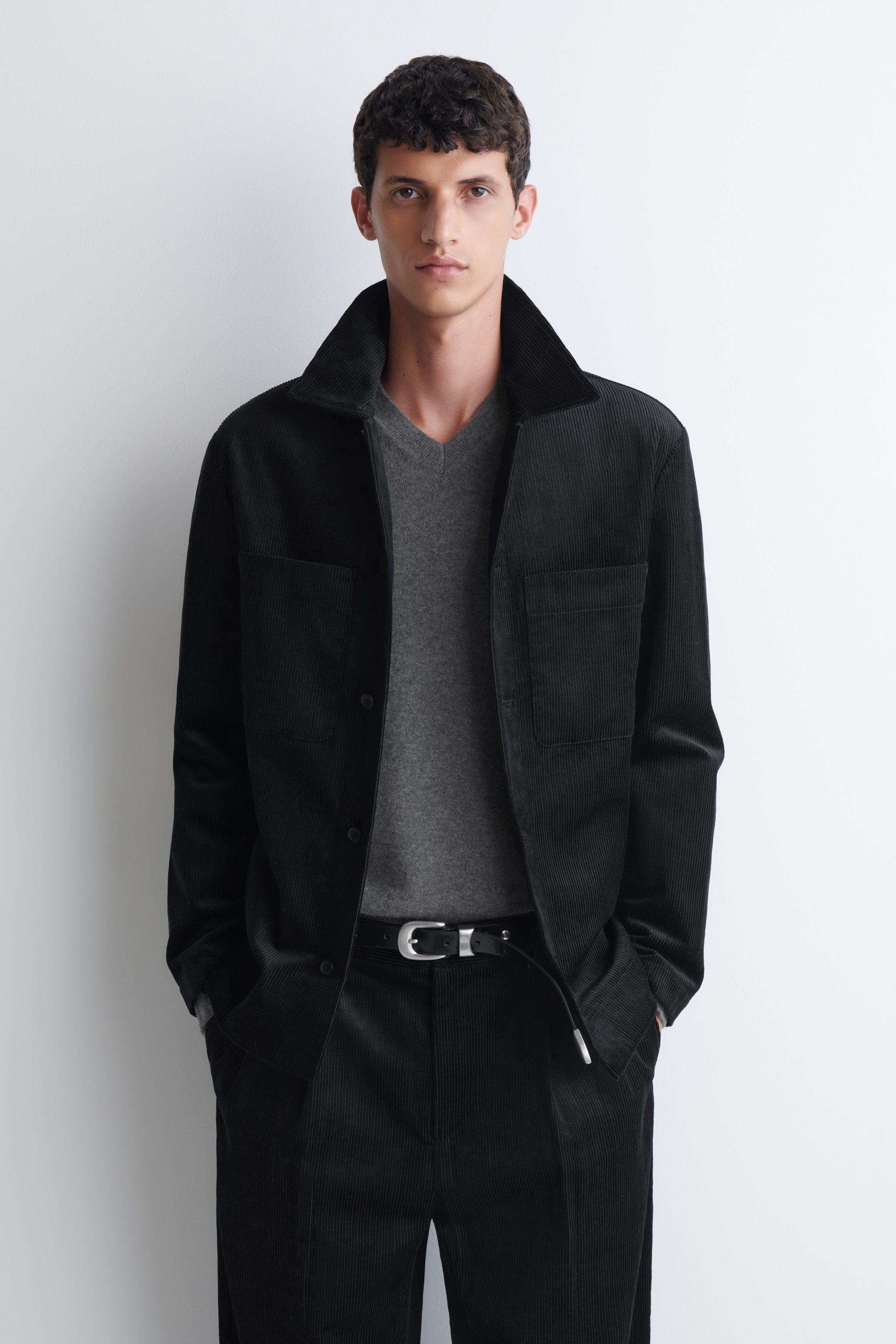Se større billede: En sort overshirt i bomuldsfløjl med spids krave og brystlommer bæres åben af modellen. Overshirten er lag-på-lag over en grå top med V-udskæring og kombineres med matchende sorte fløjlsbukser.