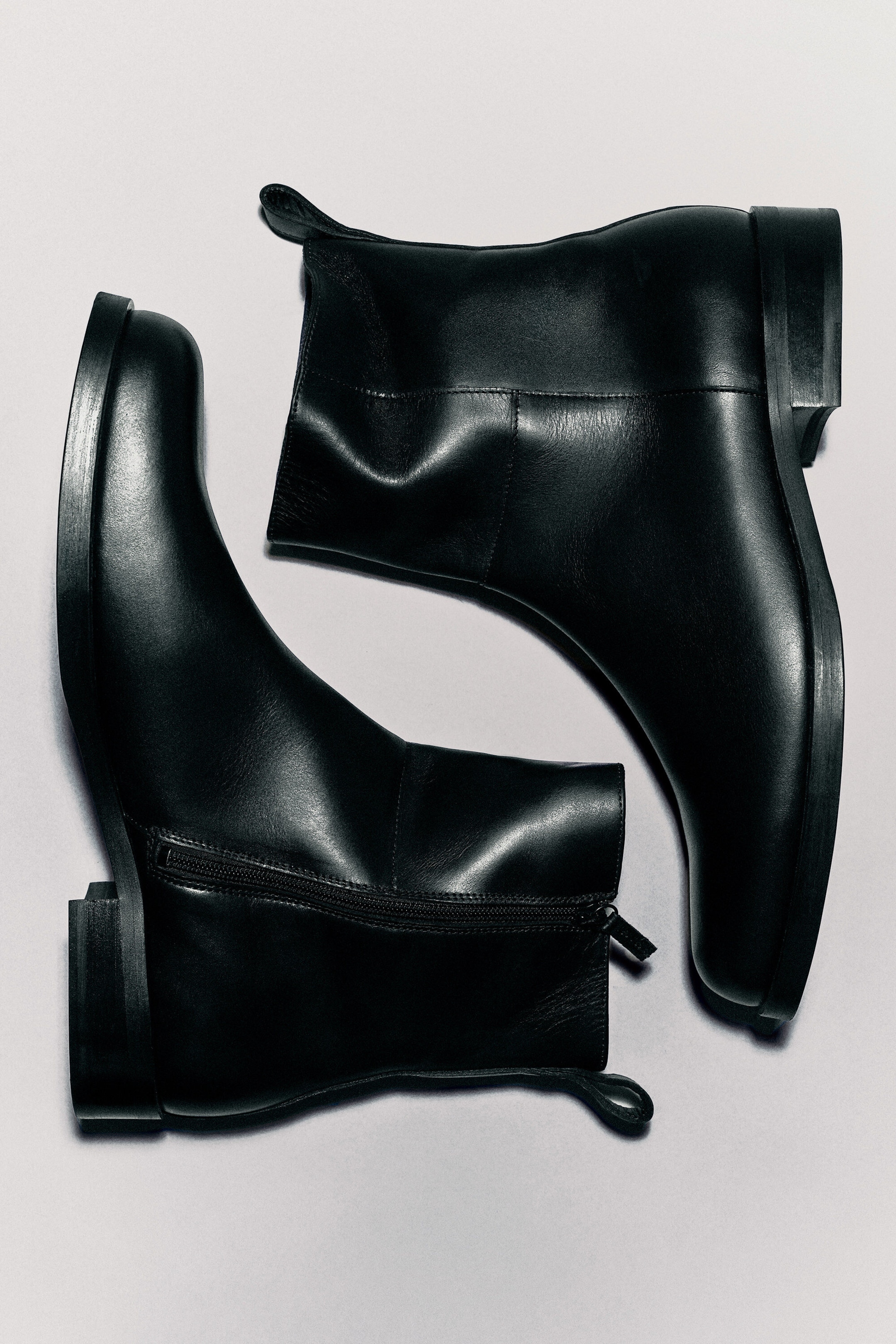 Square-toe leather ankle boots in black are laid flat in an opposing arrangement, showcasing their smooth leather construction, low block heels, and side-zip fastenings. The boots feature a rear pull tab and neat panel stitching along the shaft.