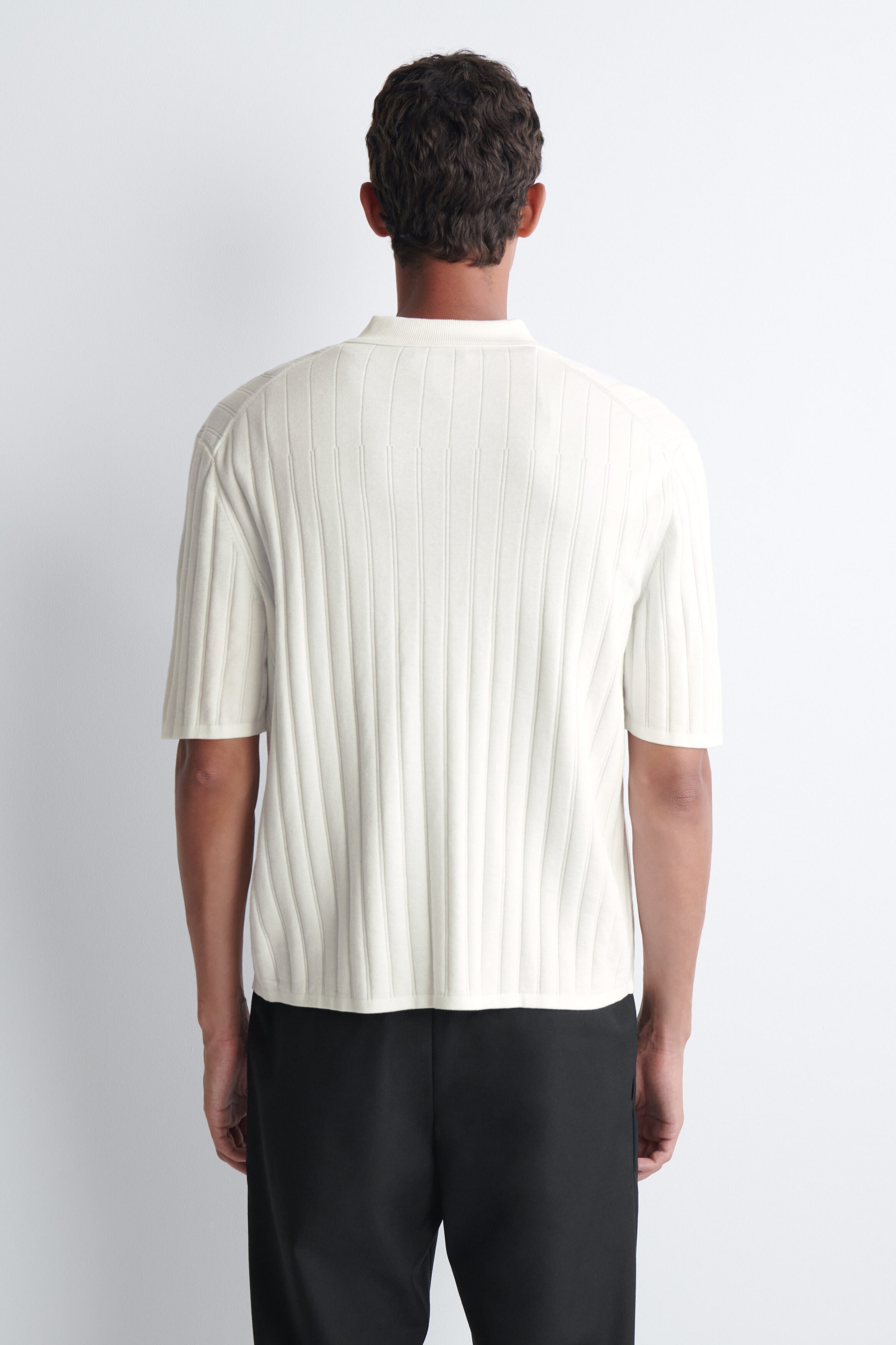 Ein gerippt gestricktes Baumwollhemd in Off-White wird vom Model getragen, von hinten gezeigt. Das kurzärmelige Hemd verfügt über vertikale Rippstruktur und einen Kragen und ist lässig über einer schwarzen Hose getragen. Ein gerippt gestricktes Baumwollhemd in Off-White wird vom Model getragen, von hinten gezeigt. Das kurzärmelige Hemd verfügt über vertikale Rippstruktur und einen Kragen und ist lässig über einer schwarzen Hose getragen.