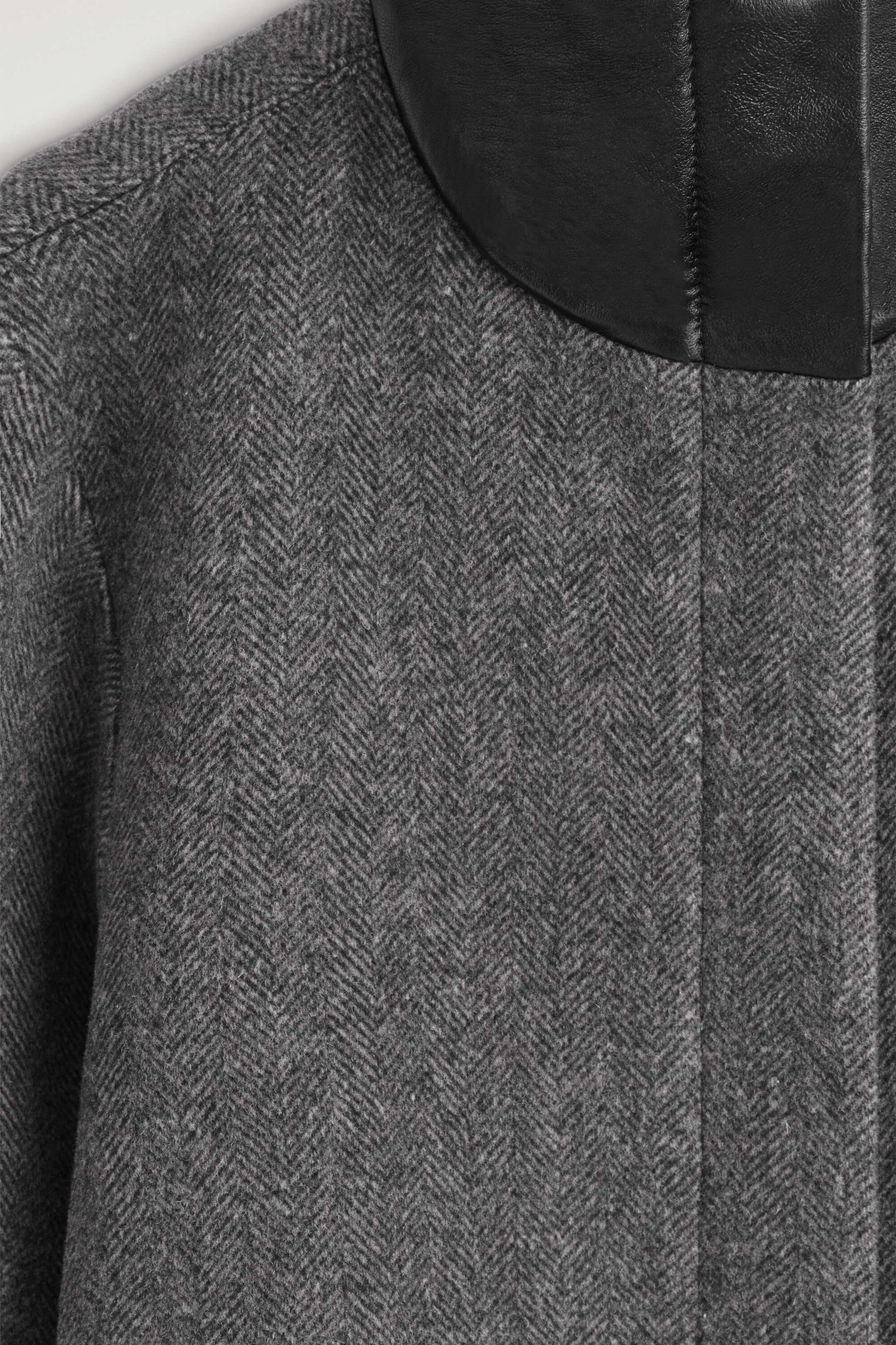 Eine Nahaufnahme der Vorderansicht einer grauen Jacke aus Wollmischgewebe im Fischgrätmuster, die ihr strukturiertes Muster im Kontrast zu einem glatten schwarzen Funnel-Neck-Kragen aus Leder zeigt. Die Aufnahme konzentriert sich auf den oberen Brust- und Schulterbereich und hebt die haptische Qualität des Stoffes hervor. Eine Nahaufnahme der Vorderansicht einer grauen Jacke aus Wollmischgewebe im Fischgrätmuster, die ihr strukturiertes Muster im Kontrast zu einem glatten schwarzen Funnel-Neck-Kragen aus Leder zeigt. Die Aufnahme konzentriert sich auf den oberen Brust- und Schulterbereich und hebt die haptische Qualität des Stoffes hervor.