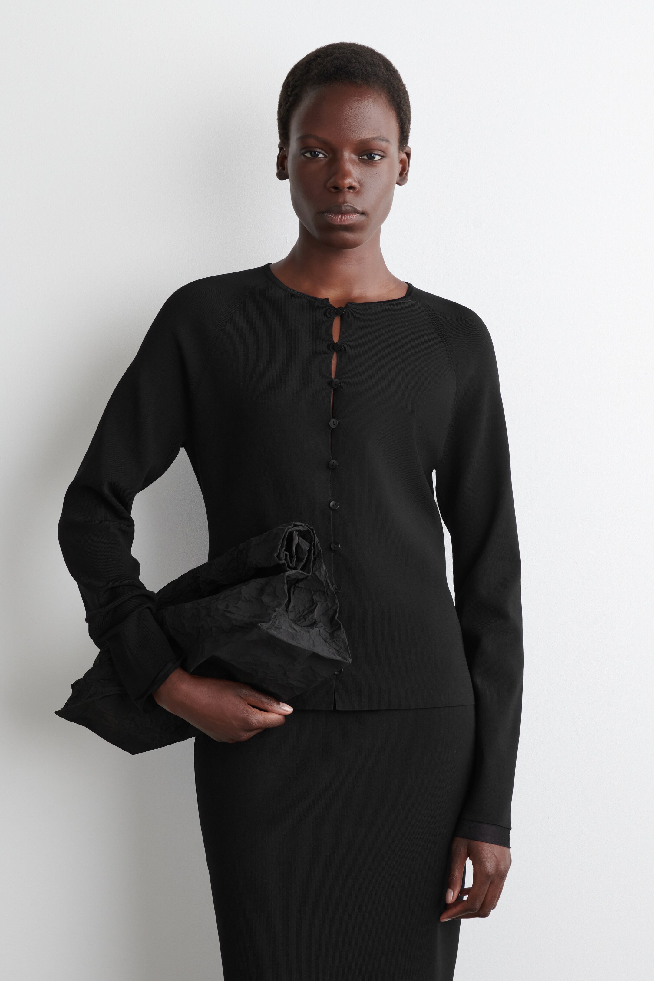 View larger image: A black cardigan is being presented front-facing, featuring a round neckline, long raglan sleeves, and a front button closure with small, dark buttons.