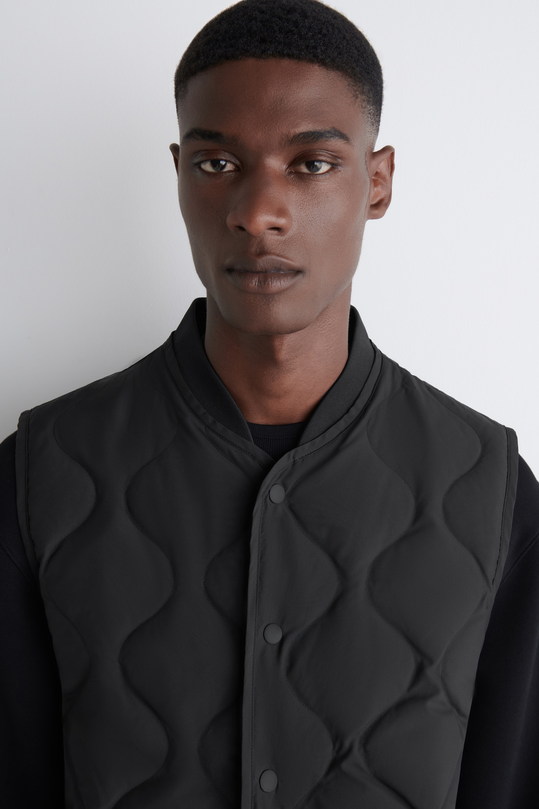 Größeres Bild anzeigen: Ein schwarzes Gilet mit einem wellenförmigen Steppmuster und Druckknopfverschlüssen wird vom Model über einem schwarzen langärmeligen Oberteil getragen. Das Gilet hat einen gerippten Kragen.