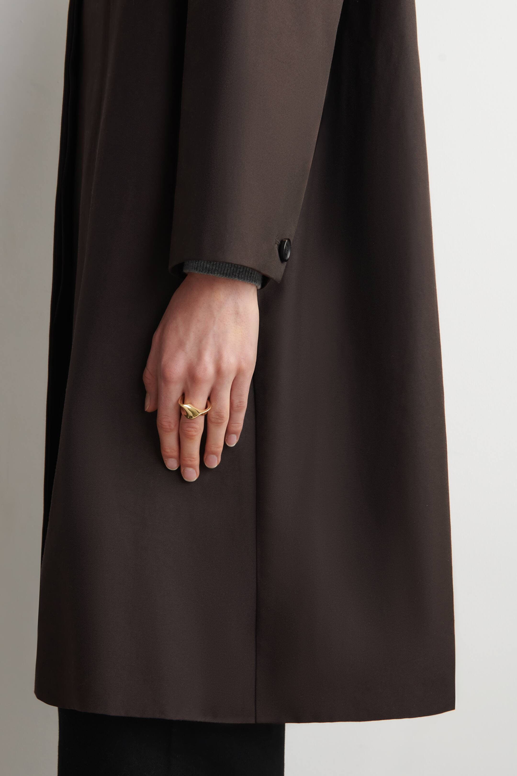 Agrandir l'image: Un manteau marron foncé présente son panneau latéral et frontal, avec sa manche longue ornée d'un unique bouton sombre près du poignet. Un poignet côtelé gris s'étend visiblement sous la manche du manteau.