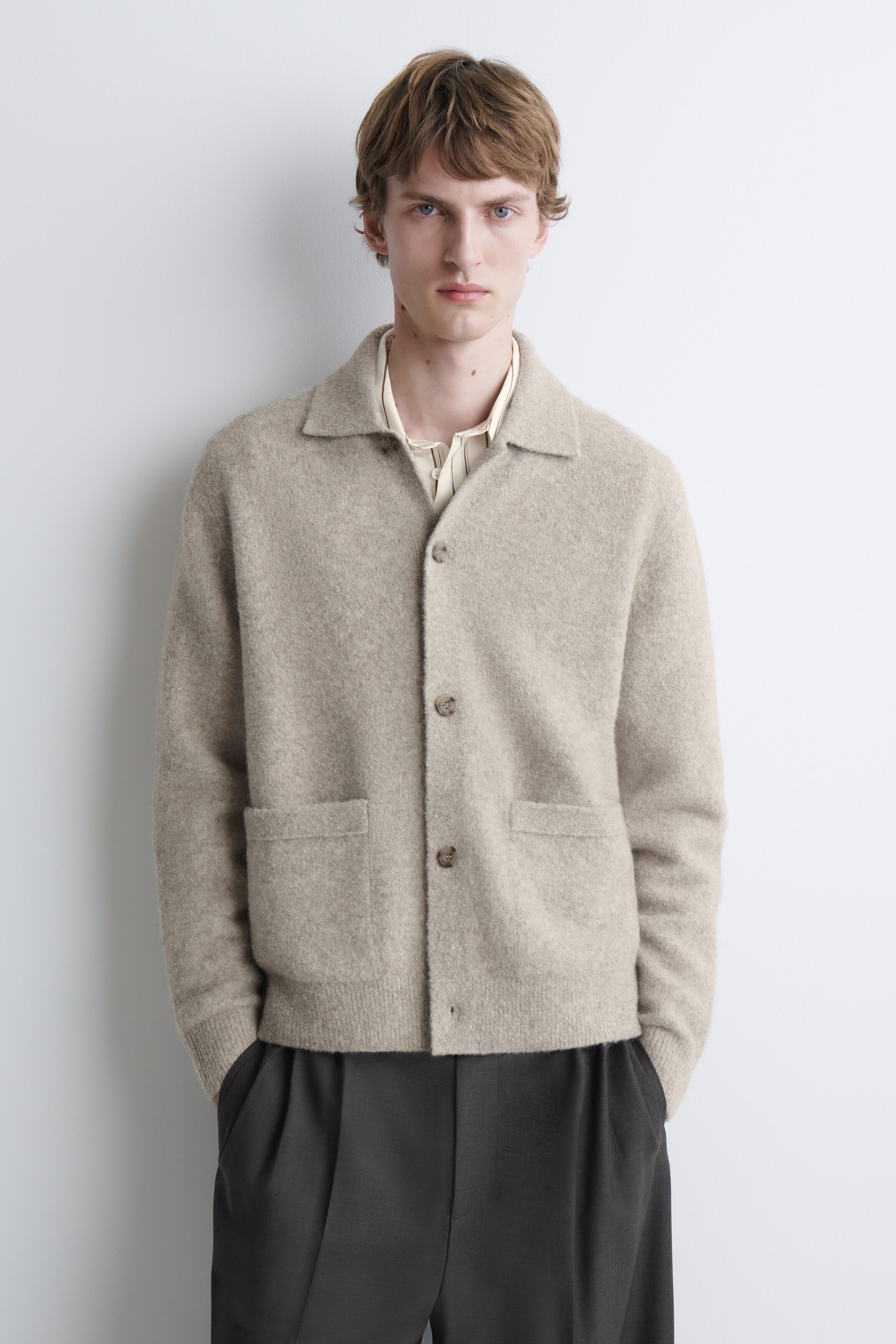 A beige brushed-knit cardigan is worn by the model, featuring a classic collar, button closure, and two front patch pockets. The cardigan is layered over a light-coloured collared shirt and paired with dark pleated trousers. A beige brushed-knit cardigan is worn by the model, featuring a classic collar, button closure, and two front patch pockets. The cardigan is layered over a light-coloured collared shirt and paired with dark pleated trousers.