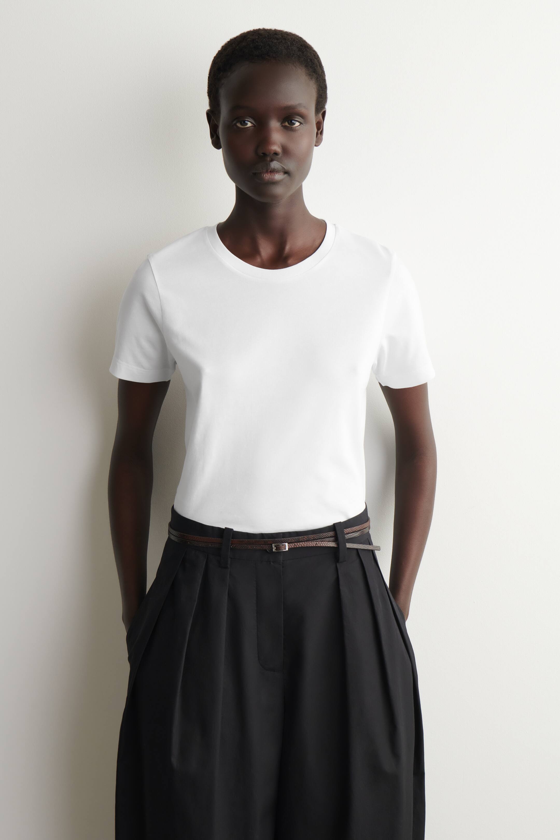 Größeres Bild ansehen: Ein weißes T-Shirt mit Rundhalsausschnitt und kurzen Ärmeln. Es präsentiert eine glatte, taillierte Silhouette und wird in dunkle Hosen gesteckt getragen.