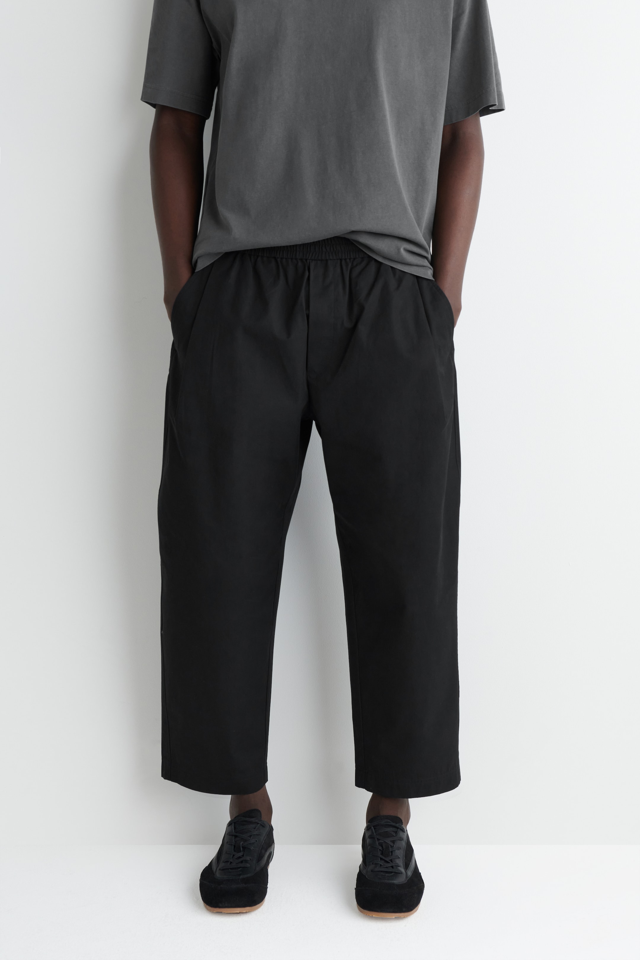 Agrandir l'image: Un pantalon noir raccourci est présenté de face, avec une taille élastiquée, des plis subtils à l'avant et des poches latérales, avec une coupe ample et décontractée se terminant au-dessus des chevilles. Ils sont stylisés avec un t-shirt gris foncé surdimensionné et des chaussures basses noires.