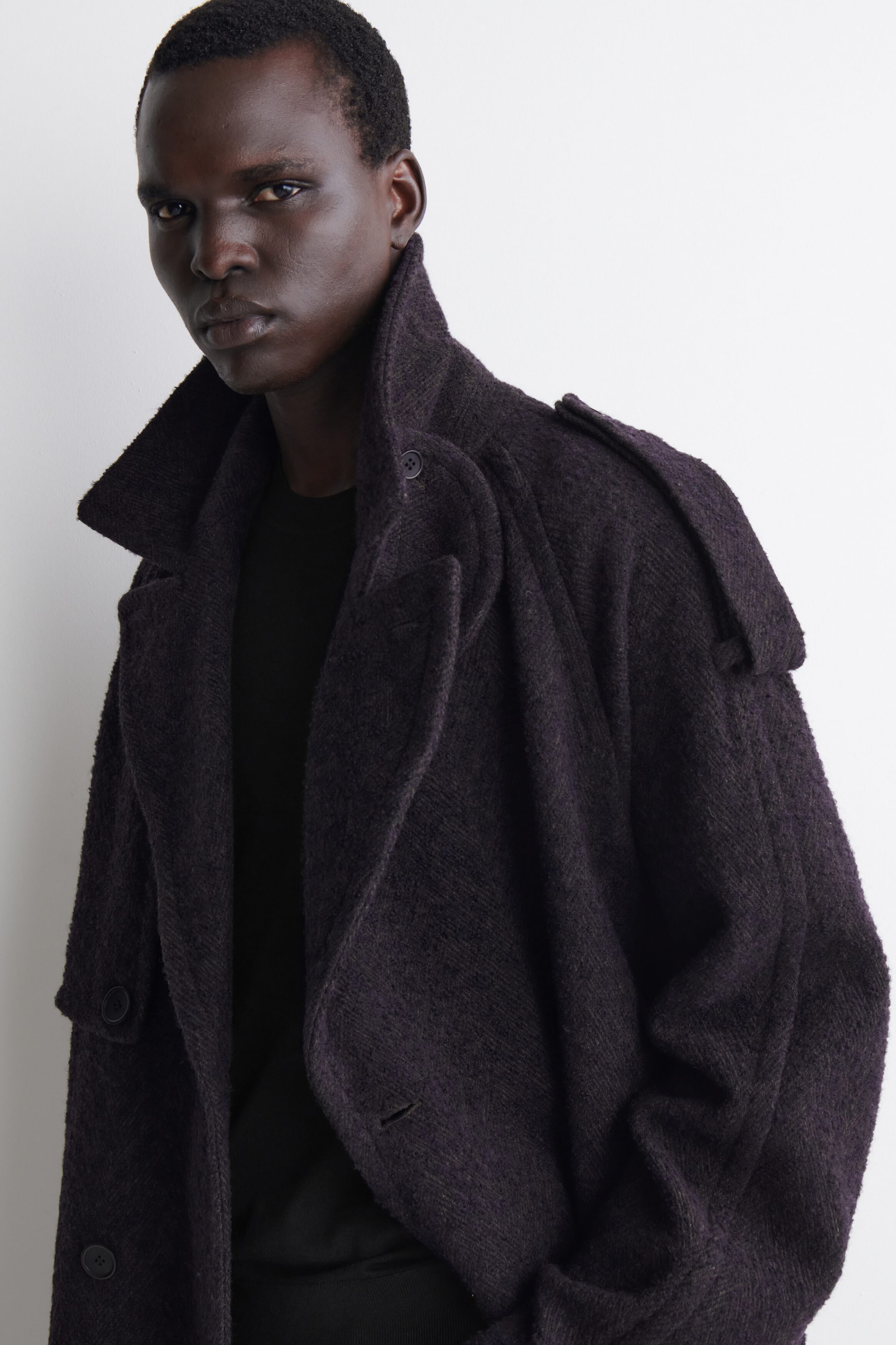 Ein Model trägt einen dunkelvioletten Trenchcoat aus strukturiertem Wollmix, in einer Nahaufnahme, die den Oberkörper und die Schulterdetails hervorhebt. Der Mantel verfügt über einen hohen, weiten Kragen, einen zweireihigen Vorderteil und eine geknöpfte Schulterklappe an der Schulter, kombiniert mit einem schwarzen Oberteil. Ein Model trägt einen dunkelvioletten Trenchcoat aus strukturiertem Wollmix, in einer Nahaufnahme, die den Oberkörper und die Schulterdetails hervorhebt. Der Mantel verfügt über einen hohen, weiten Kragen, einen zweireihigen Vorderteil und eine geknöpfte Schulterklappe an der Schulter, kombiniert mit einem schwarzen Oberteil.