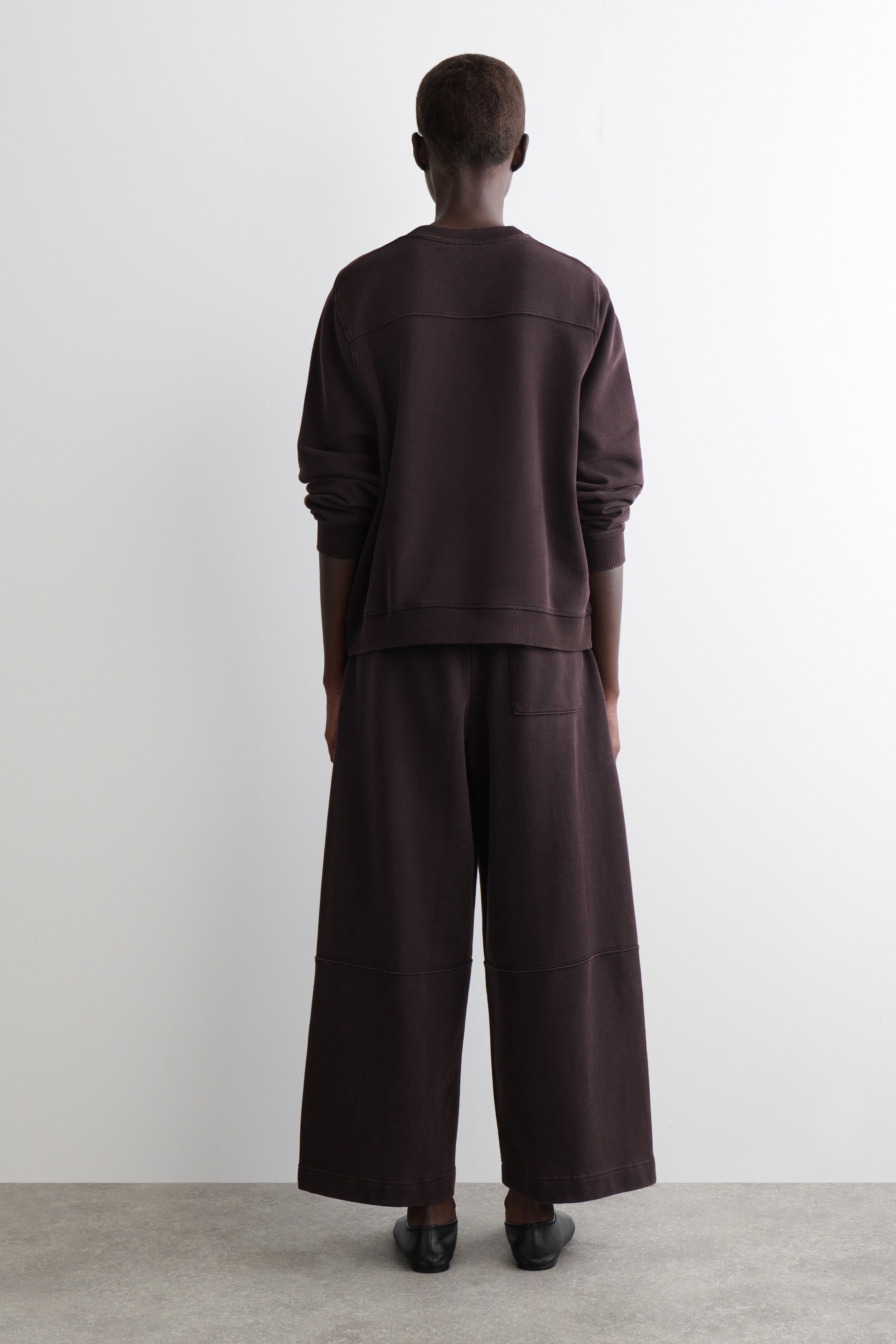 View larger image: A dark brown loungewear set is being shown from the back, featuring a long-sleeved sweatshirt with horizontal shoulder seams and a ribbed hem, and wide-leg pants with visible paneling and a back patch pocket. The pants are falling to a cropped length, and the set is presenting a relaxed fit.