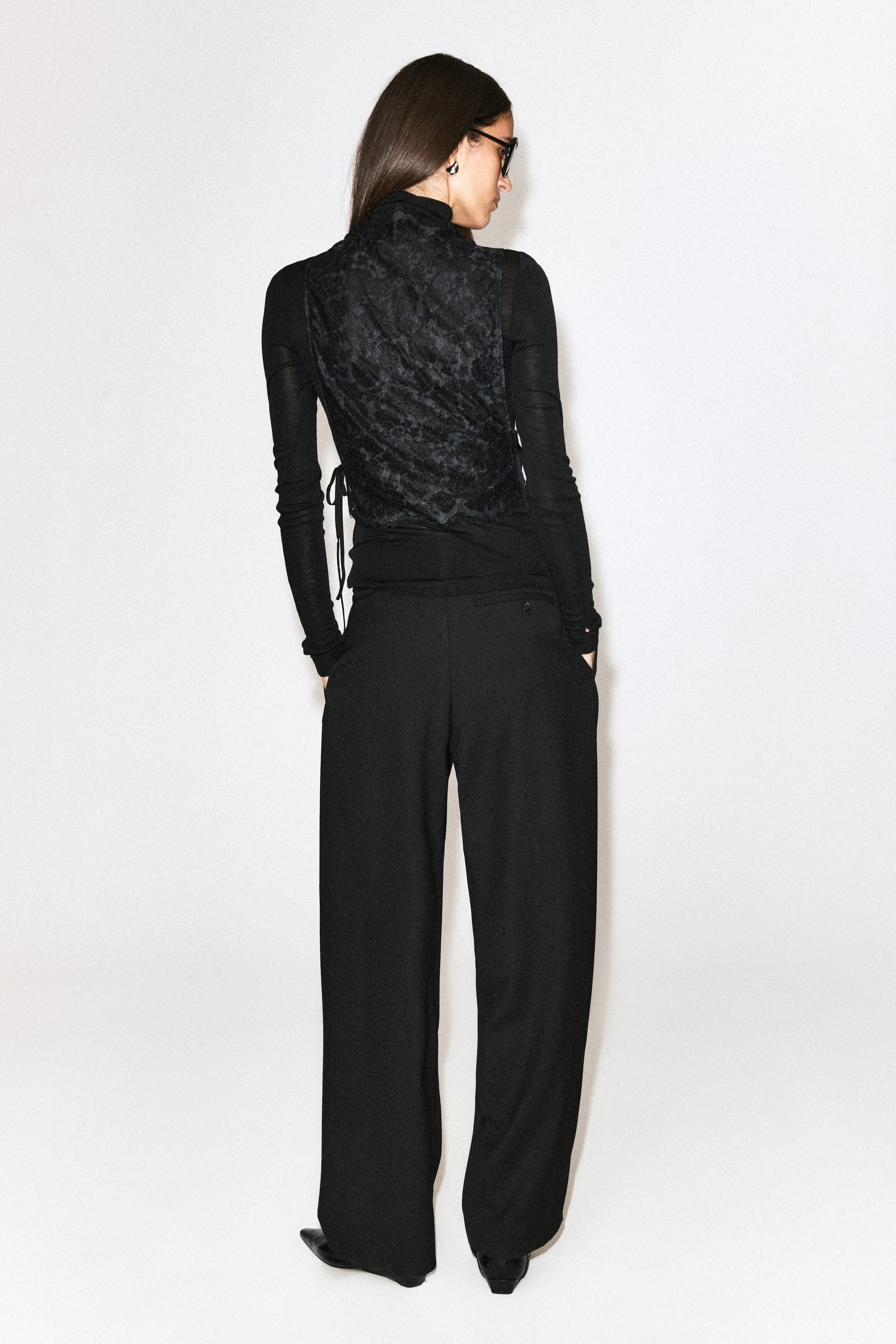 The model is wearing a black scalloped lace bib layered over a black long-sleeved top and wide-leg trousers. The bib features a high collar, a floral lace pattern, a scalloped hem, and side-tie details. The model is wearing a black scalloped lace bib layered over a black long-sleeved top and wide-leg trousers. The bib features a high collar, a floral lace pattern, a scalloped hem, and side-tie details.