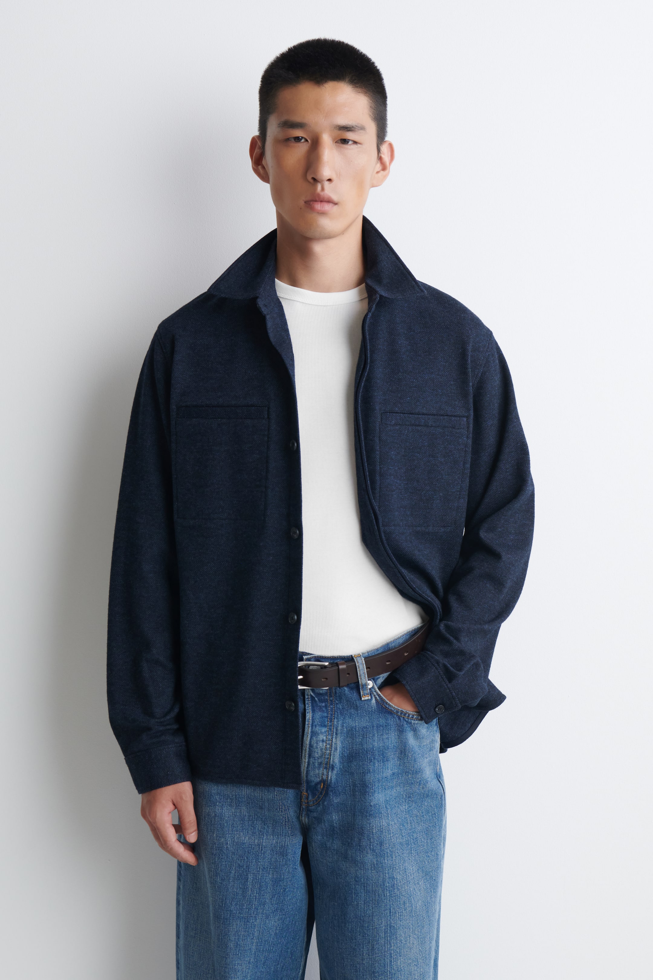 Größeres Bild anzeigen: Ein dunkelblaues Overshirt in lockerer Passform mit spitzem Kragen, zwei Brusttaschen mit Patte und langen Ärmeln mit geknöpften Manschetten, offen getragen und mit einem weißen T-Shirt darunter.