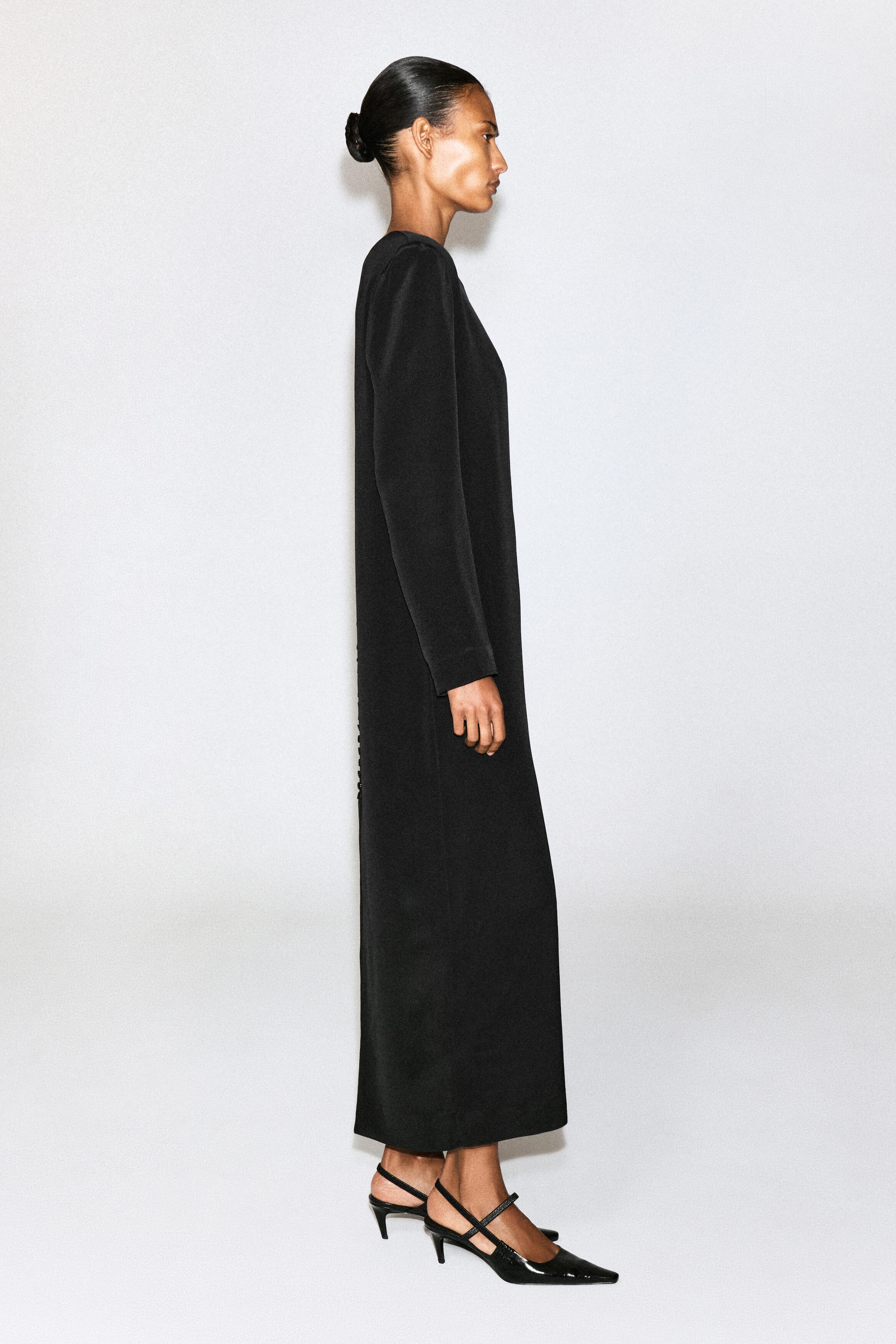 A black long-sleeved maxi dress with a straight, tailored silhouette is being worn by the model, who is standing in profile. The ankle-length dress is styled with black patent pointed-toe slingback heels.