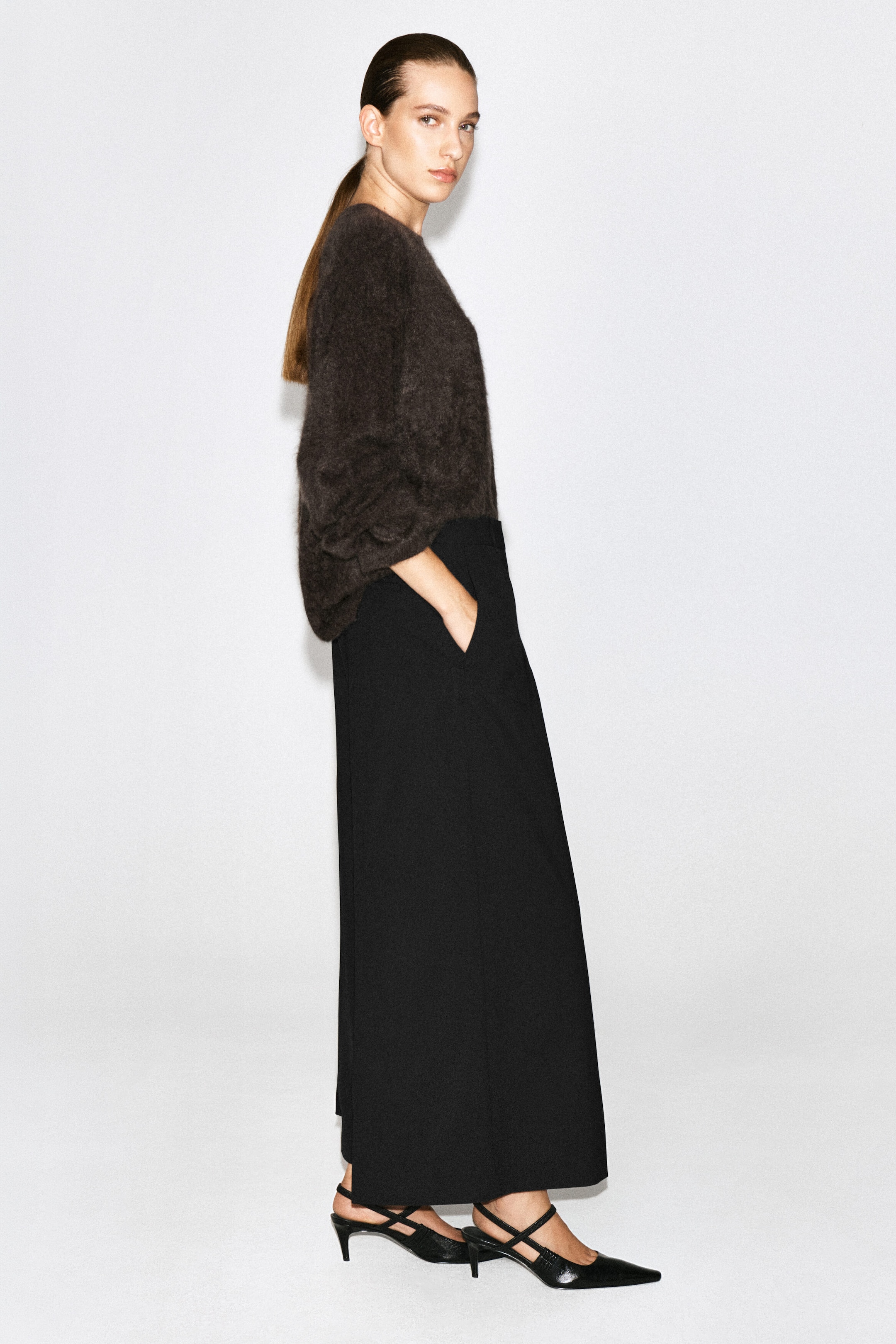 Black wool-blend wide-leg trousers are being worn by the model, who is standing in profile with one hand resting in the side pocket. The high-waisted trousers are styled with a dark, textured knit jumper tucked in at the waist and black pointed-toe slingback heels. Black wool-blend wide-leg trousers are being worn by the model, who is standing in profile with one hand resting in the side pocket. The high-waisted trousers are styled with a dark, textured knit jumper tucked in at the waist and black pointed-toe slingback heels.