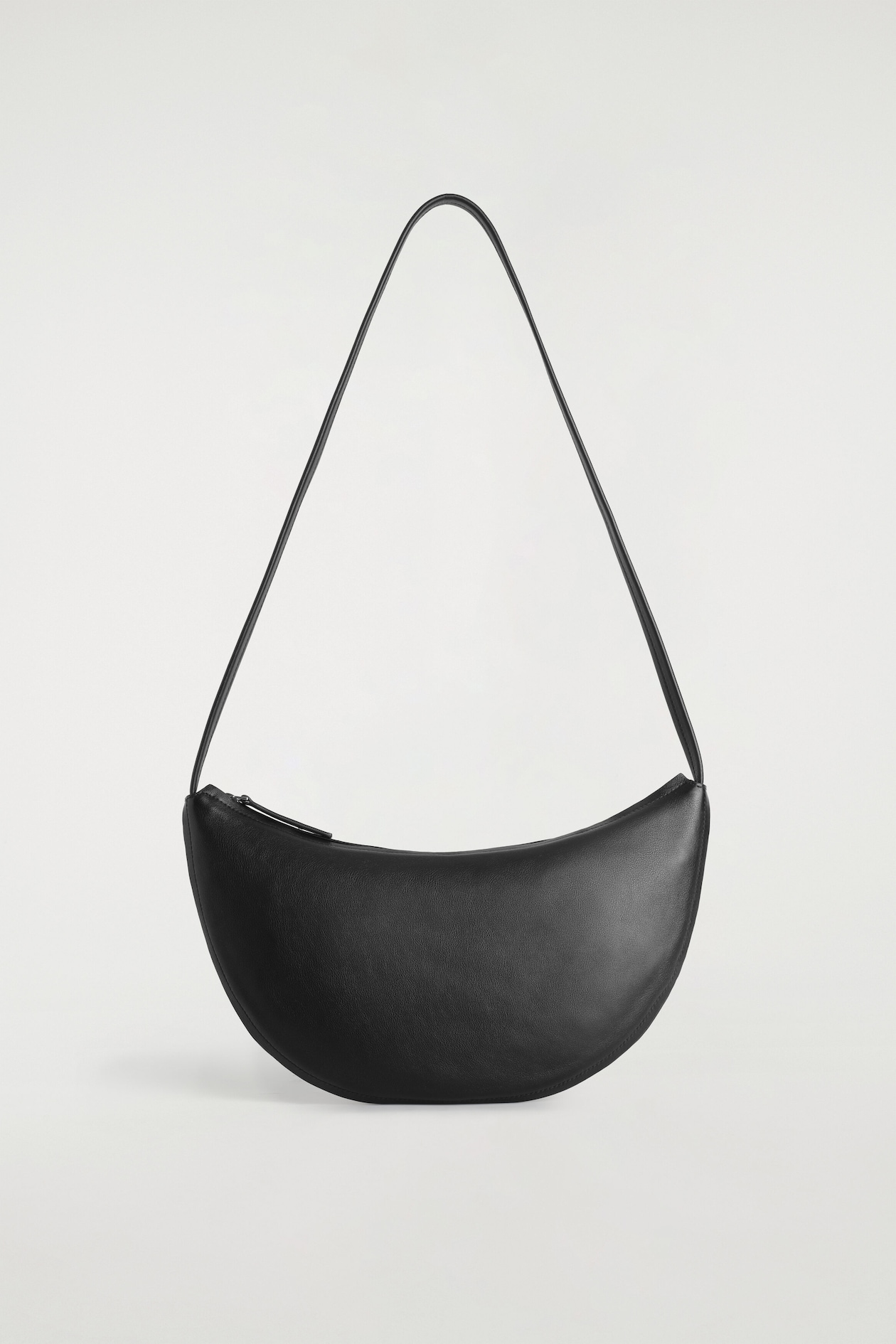 A black leather shoulder bag in a curved, crescent silhouette is standing upright, featuring a top zip closure and a long, slim shoulder strap.