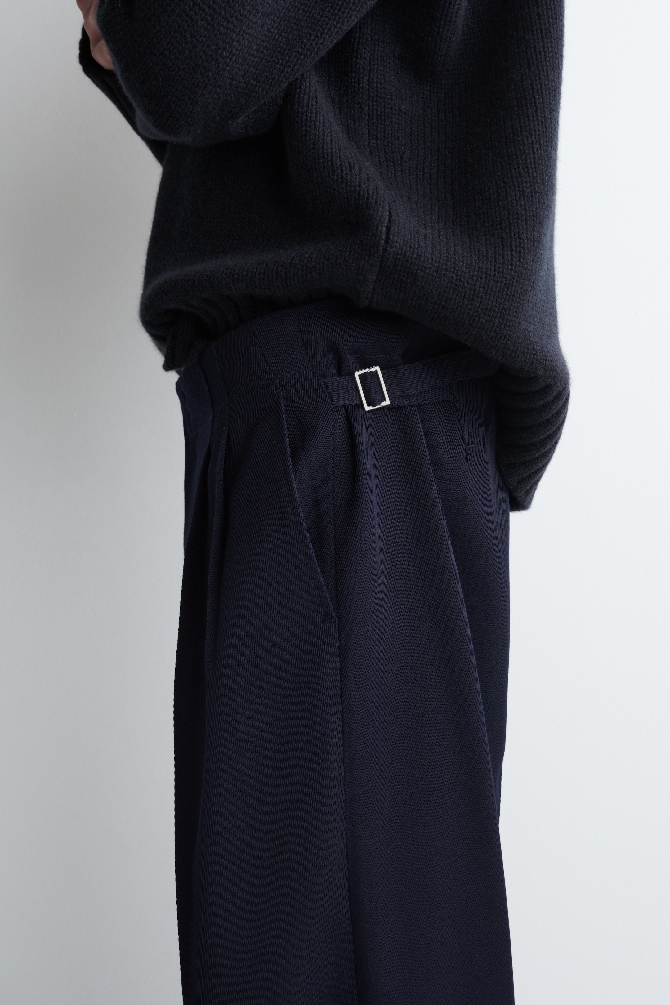 Agrandir l'image: Un pantalon plissé bleu foncé est présenté de profil, avec des plis à l'avant, un ajusteur latéral avec une boucle en argent à la taille, et une poche latérale. Un pull en tricot bleu foncé avec un ourlet côtelé est porté par-dessus le pantalon, cachant partiellement la ceinture.