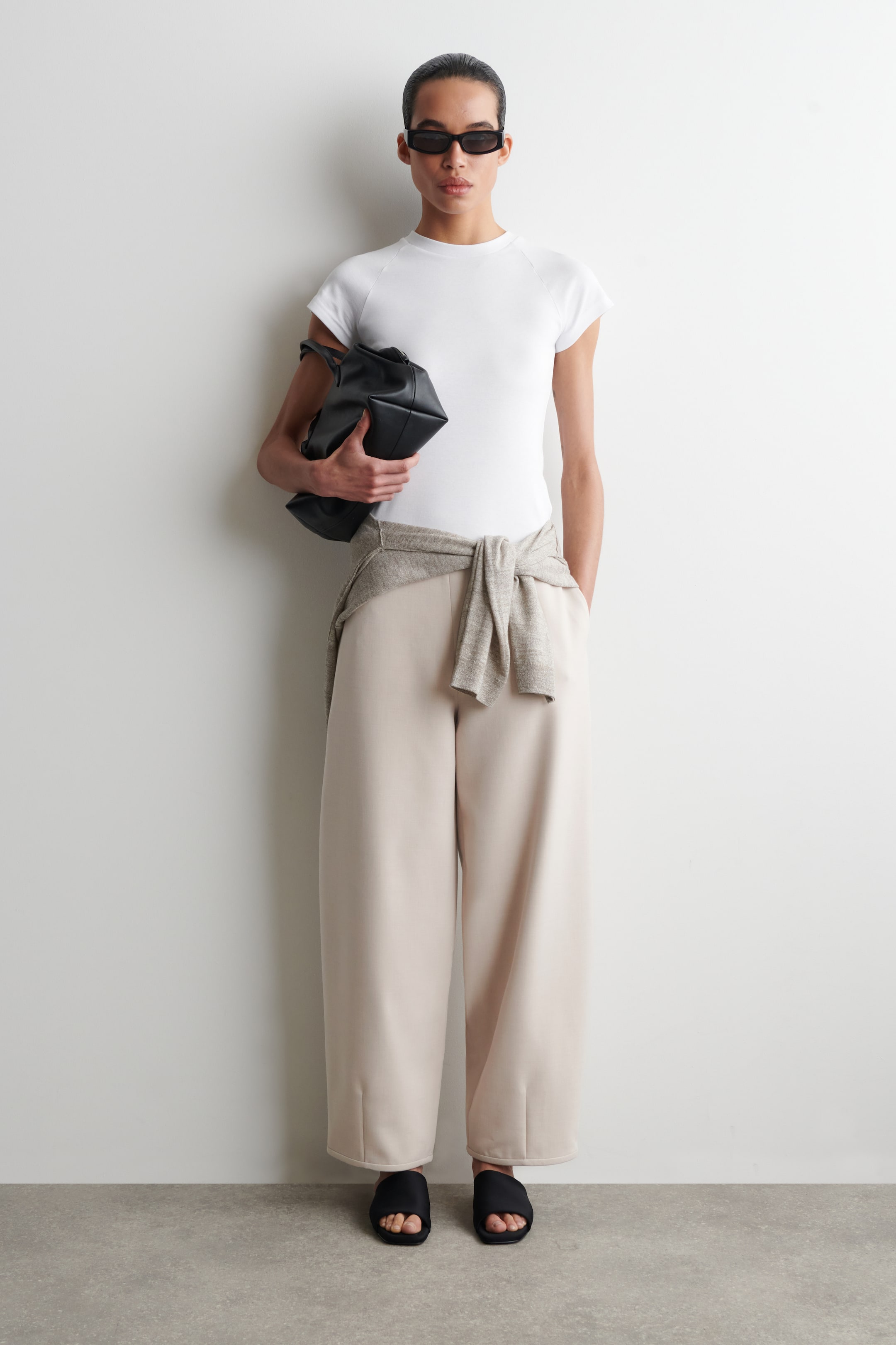 Se større billede: De lyse beige bukser præsenteres forfra og har en afslappet, tøndeformet silhuet lavet af et glat vævet stof. En lysegrå sweater er bundet omkring taljen og dækker linningen på de cropped bukser.