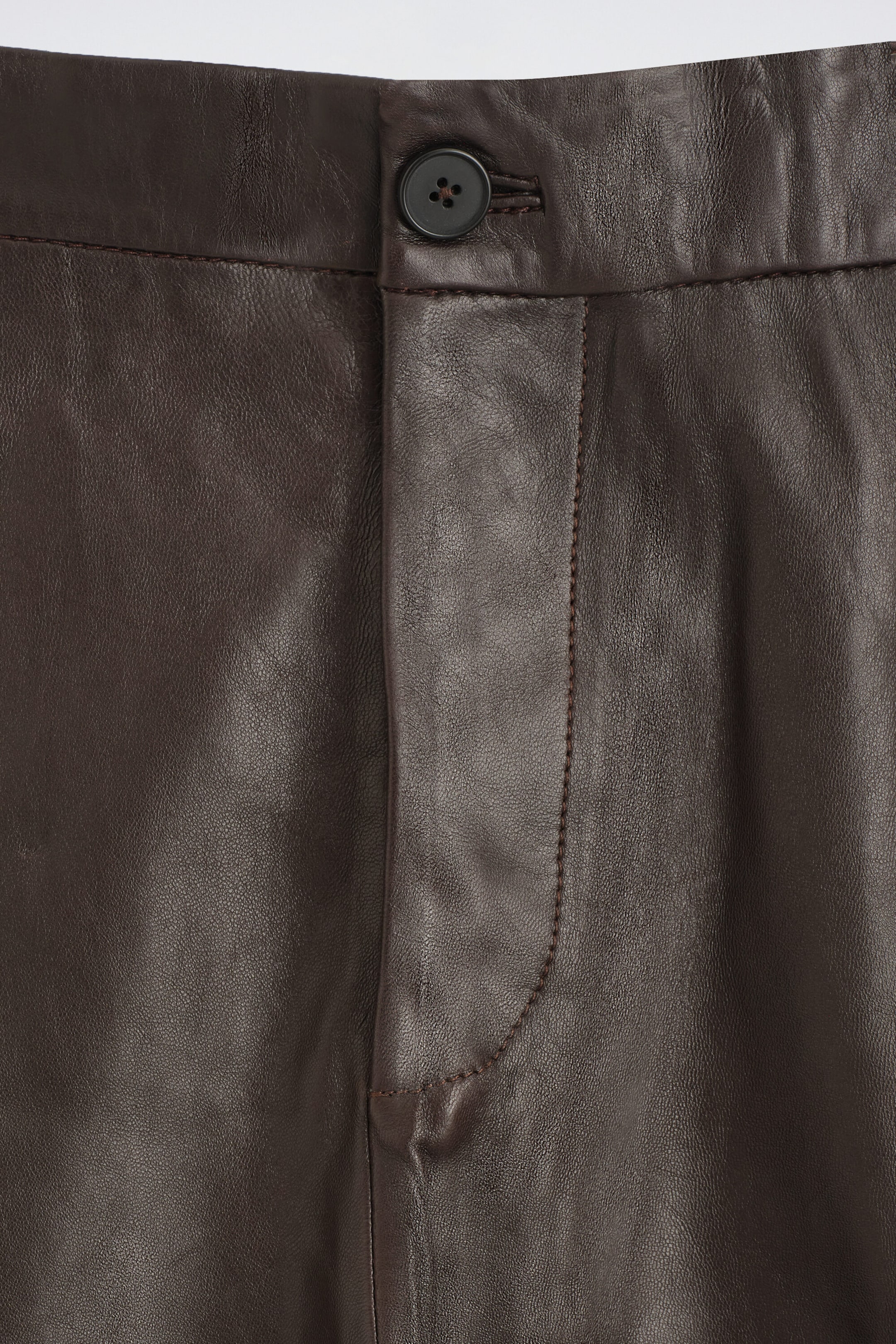 These dark brown leather pants are shown in a close-up front view of the waistband, featuring a single dark button closure and a stitched fly cover that highlights the leather’s natural grain. These dark brown leather pants are shown in a close-up front view of the waistband, featuring a single dark button closure and a stitched fly cover that highlights the leather’s natural grain.