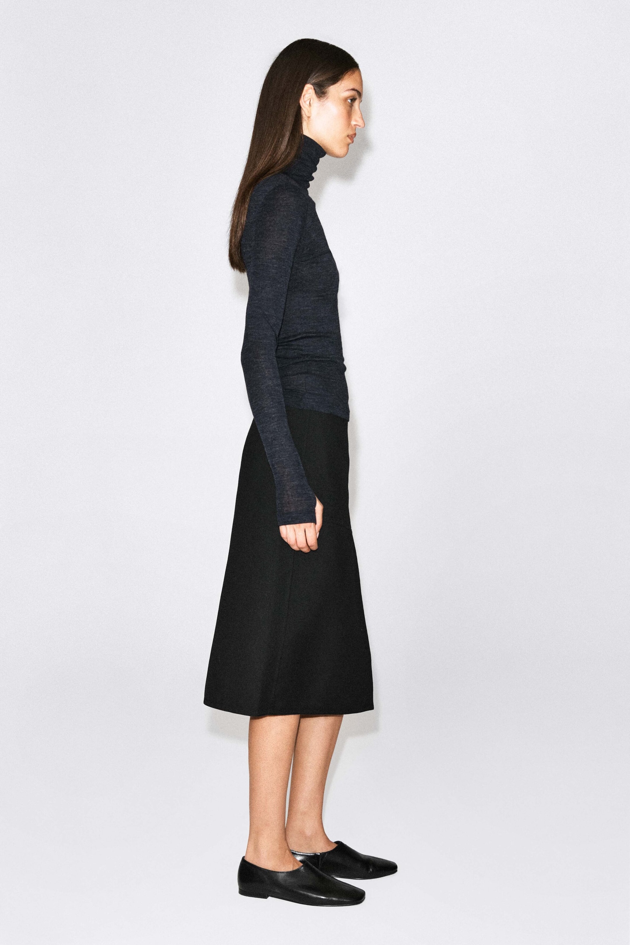 A dark gray merino wool high-neck top is being worn by the model, who is standing in profile. The fitted, long-sleeved top is styled with a black mid-length skirt and black flat leather shoes. A dark gray merino wool high-neck top is being worn by the model, who is standing in profile. The fitted, long-sleeved top is styled with a black mid-length skirt and black flat leather shoes.