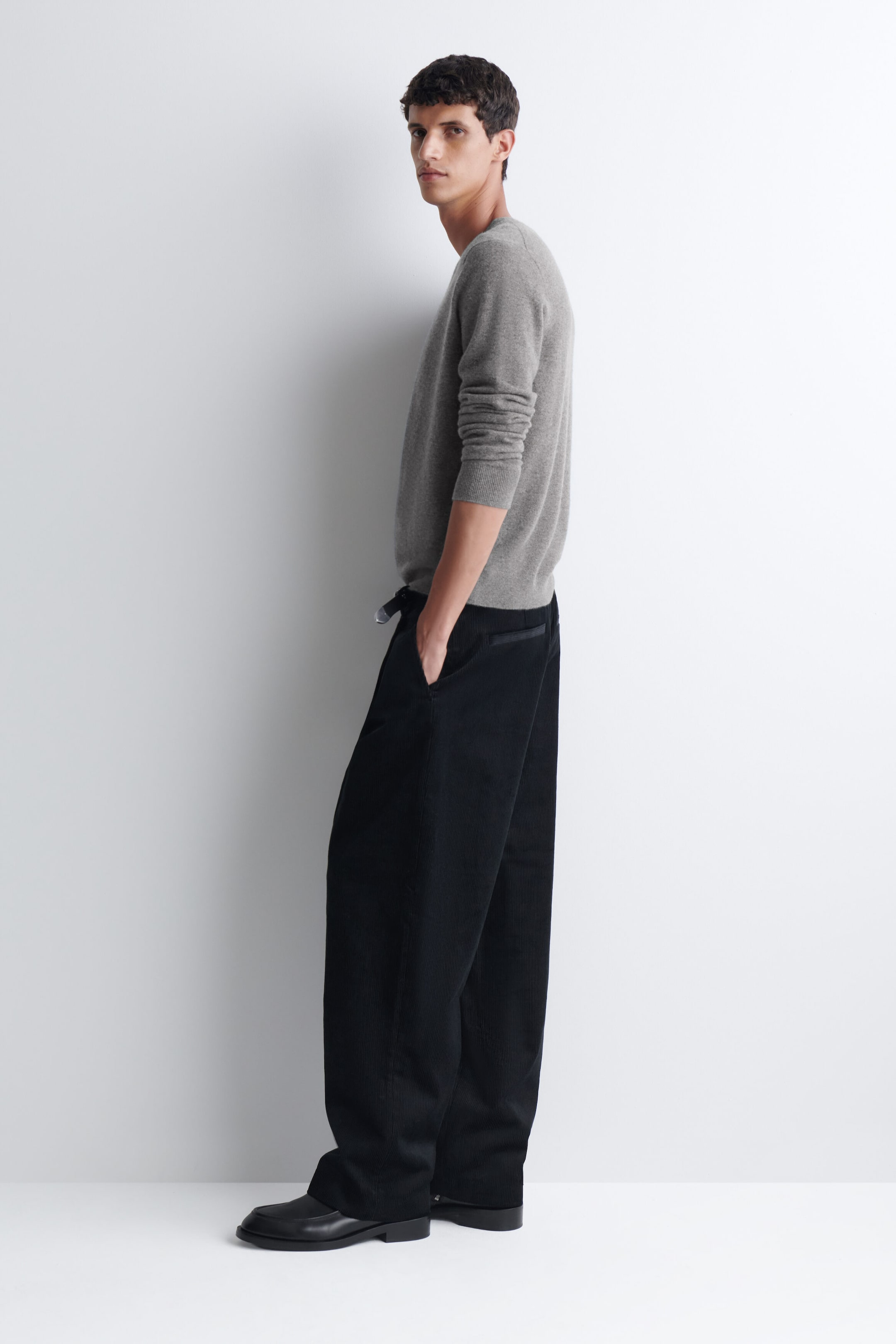 Pleated relaxed-fit tapered corduroy pants in black are being worn by the model, who is standing with one hand resting in the side pocket. The pants are styled with a long-sleeved grey crewneck sweater and black leather shoes. Pleated relaxed-fit tapered corduroy pants in black are being worn by the model, who is standing with one hand resting in the side pocket. The pants are styled with a long-sleeved grey crewneck sweater and black leather shoes.