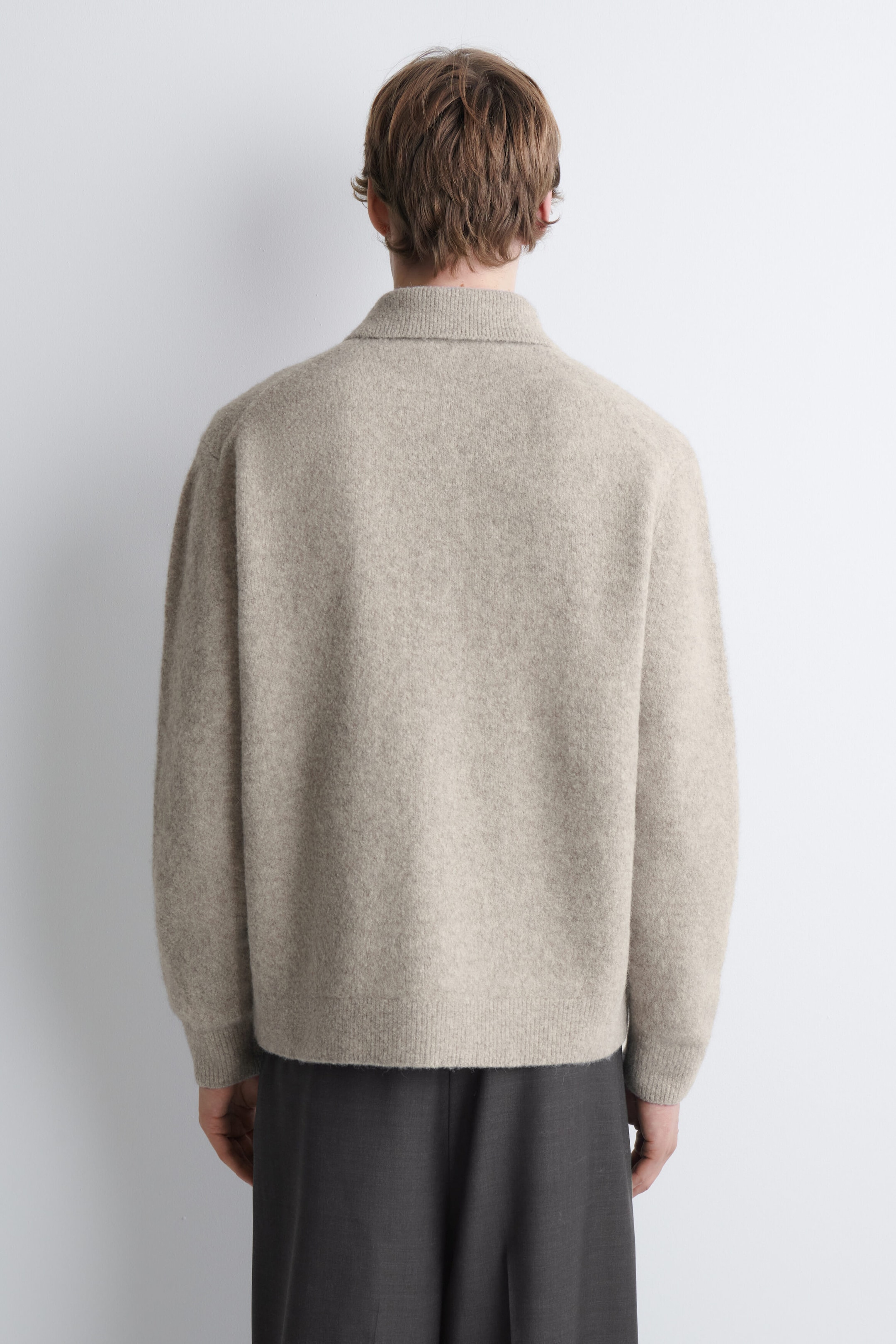 A beige brushed-knit cardigan featuring a classic collar and ribbed trims is being worn by the model, shown from behind. The cardigan is paired with dark grey trousers. A beige brushed-knit cardigan featuring a classic collar and ribbed trims is being worn by the model, shown from behind. The cardigan is paired with dark grey trousers.