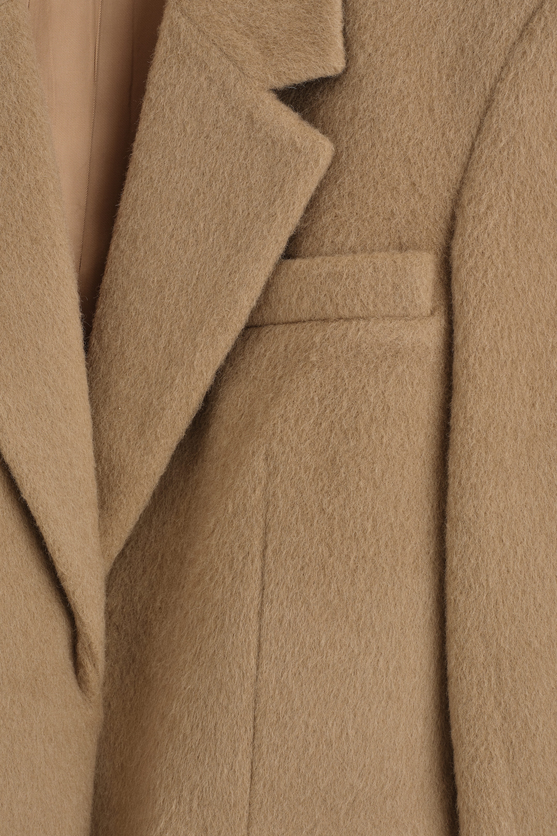 A camel-colored brushed wool blazer is shown in a close-up front view, highlighting its textured, fuzzy fabric. The detail shot emphasizes a sharp notch lapel, a welt chest pocket, and a smooth, color-matched inner lining. A camel-colored brushed wool blazer is shown in a close-up front view, highlighting its textured, fuzzy fabric. The detail shot emphasizes a sharp notch lapel, a welt chest pocket, and a smooth, color-matched inner lining.