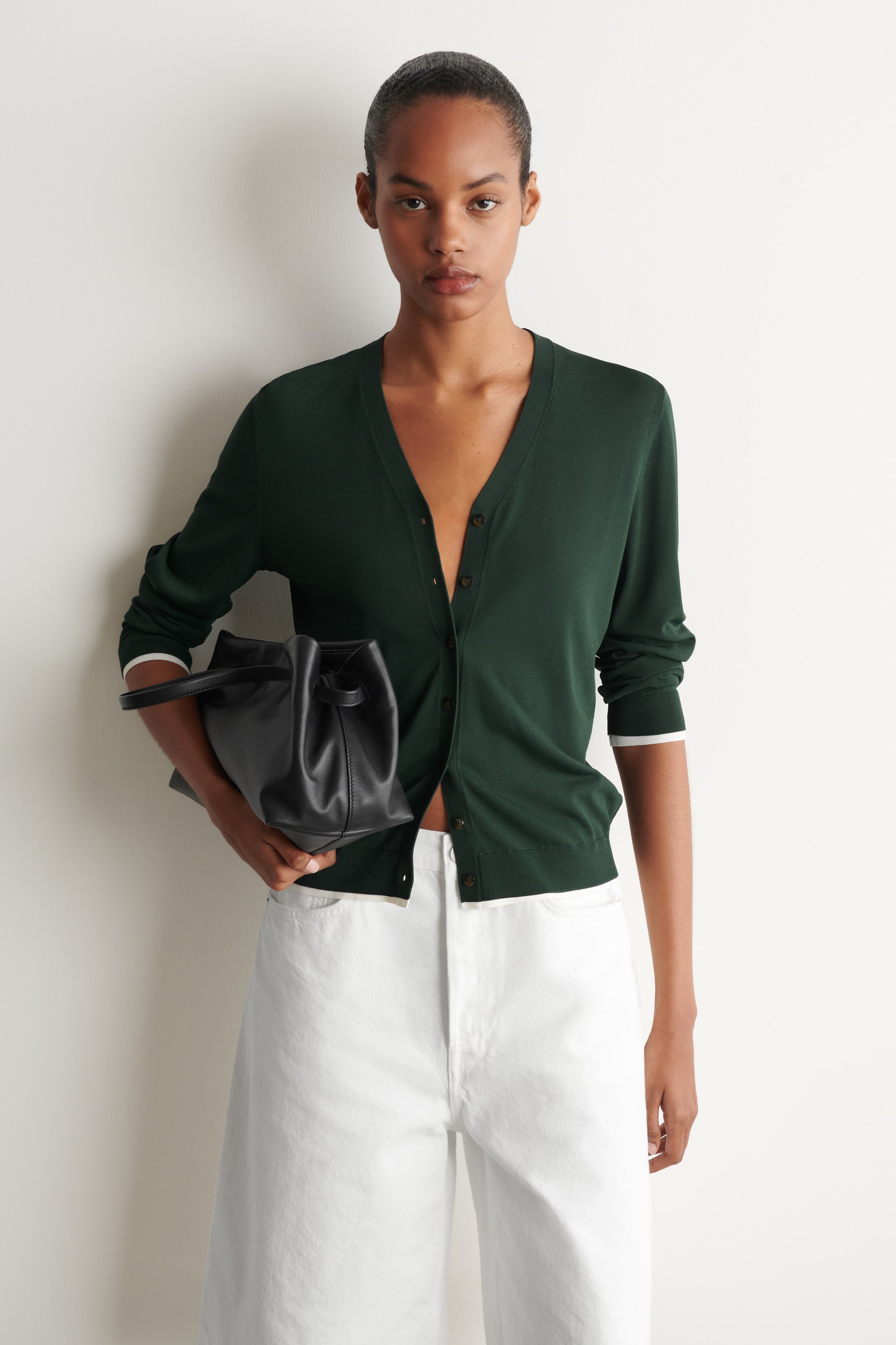 View larger image: An evergreen knitted cardigan, featuring a V-neck and contrasting white trim on its cuffs and hem, is being worn partially unbuttoned. The model is wearing it with white trousers and holding a black handbag.