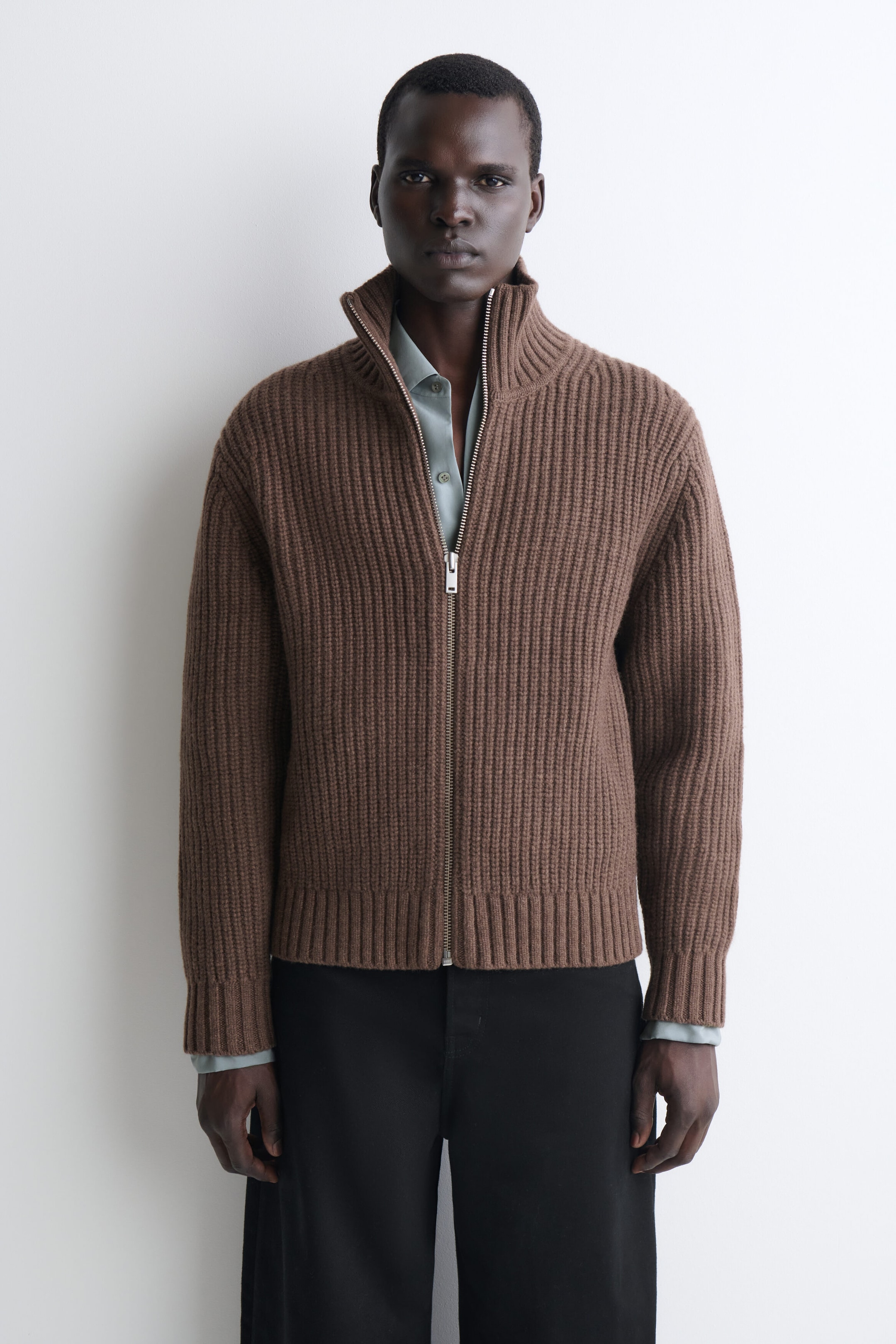 A brown rib-knit zip-up cardigan is shown from the front, featuring a high stand collar, a silver-tone front zipper, and ribbed cuffs and hem. The cardigan is styled over a light grayish-blue button-down shirt and black trousers, highlighting its relaxed, structured fit and heavyweight texture. A brown rib-knit zip-up cardigan is shown from the front, featuring a high stand collar, a silver-tone front zipper, and ribbed cuffs and hem. The cardigan is styled over a light grayish-blue button-down shirt and black trousers, highlighting its relaxed, structured fit and heavyweight texture.
