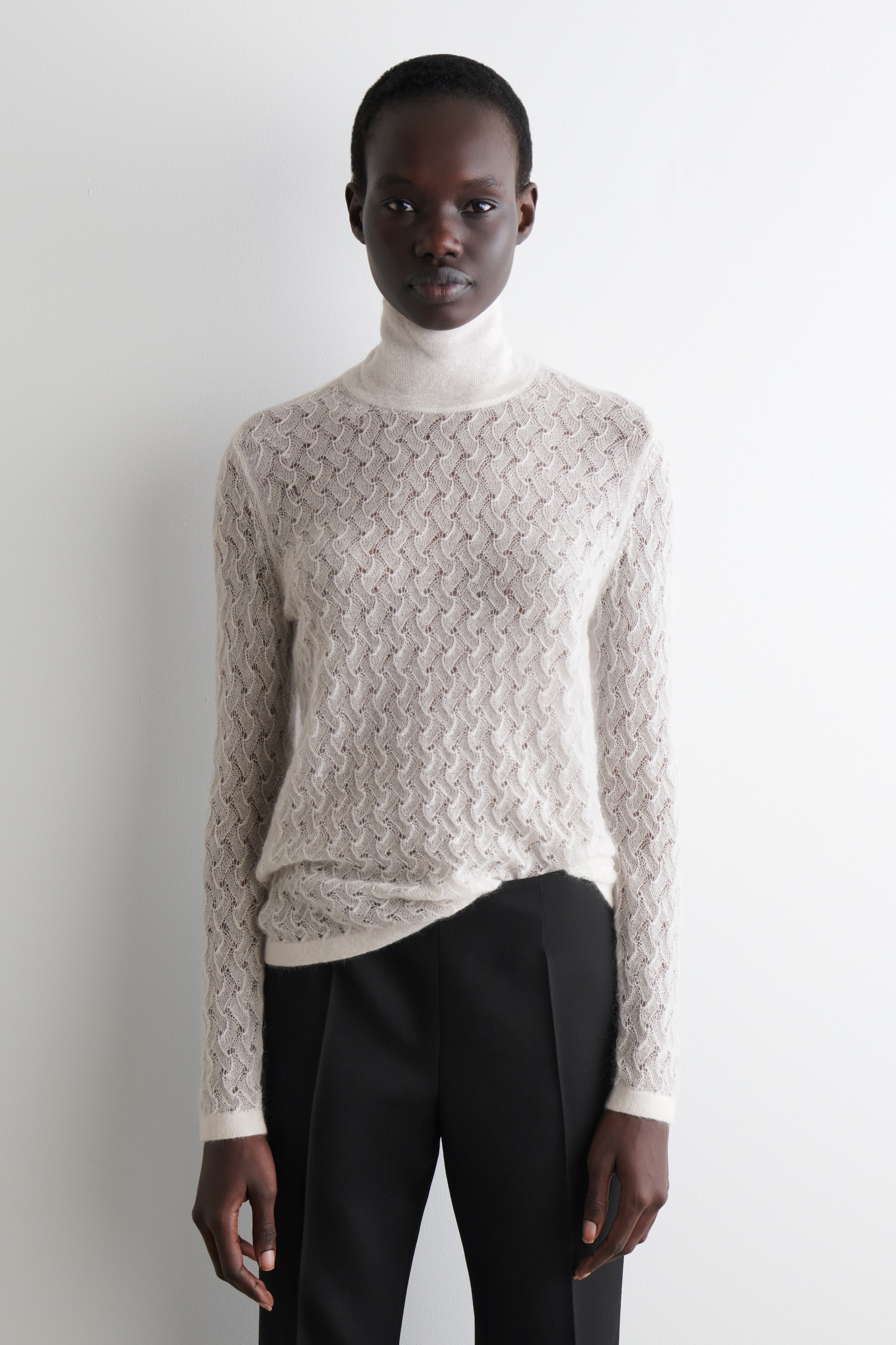 Visa större bild: En ljusbeige långärmad topp presenteras framifrån, med ett delikat, subtilt genomskinligt pointelle-stickmönster över dess kropp och ärmar. Den har en ribbad trattkrage, ribbade manschetter och en ribbad fåll, och sitter slimmat.