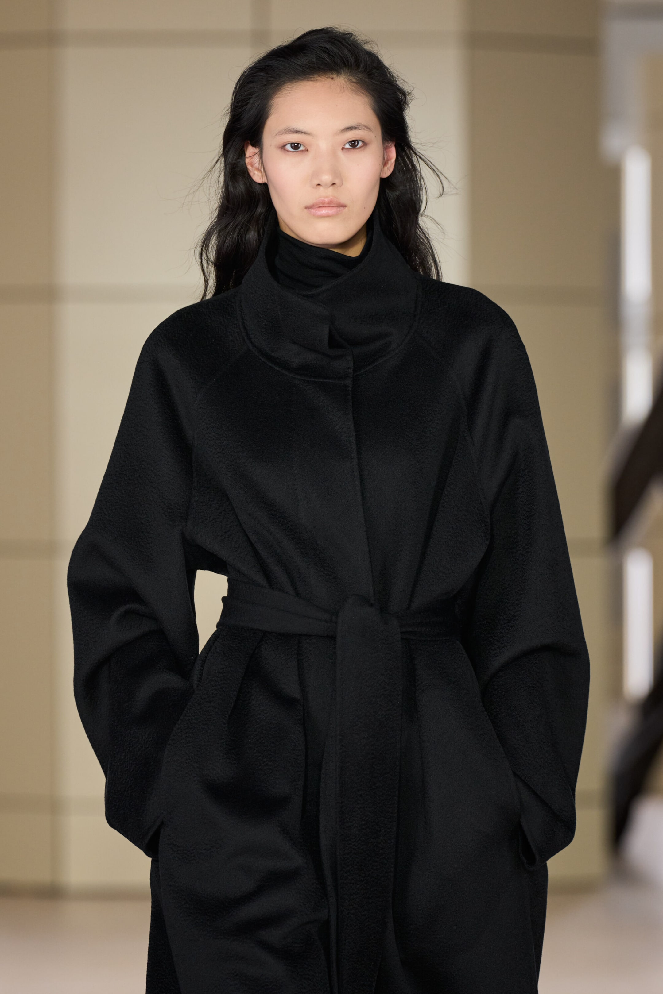 A black belted double-faced cashmere coat is shown from the front, featuring a high, structured funnel neck, relaxed raglan sleeves, and a matching self-tie belt cinching the waist. The coat is layered over a black roll-neck, showcasing its soft, textured fabric and relaxed silhouette.