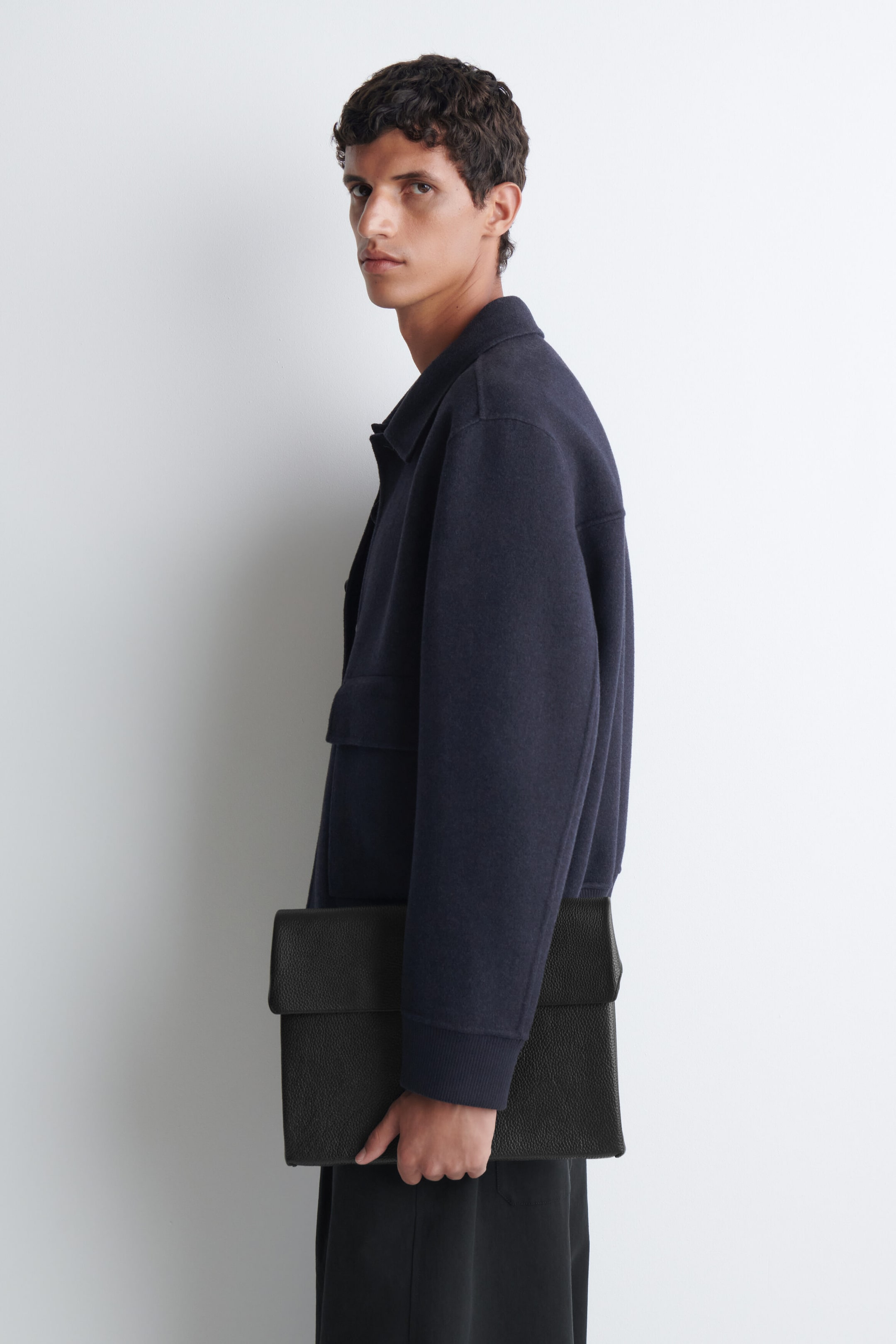 The black leather envelope laptop sleeve, featuring a textured pebbled finish and a fold-over top flap, is being carried by the model at their side. The black leather envelope laptop sleeve, featuring a textured pebbled finish and a fold-over top flap, is being carried by the model at their side.