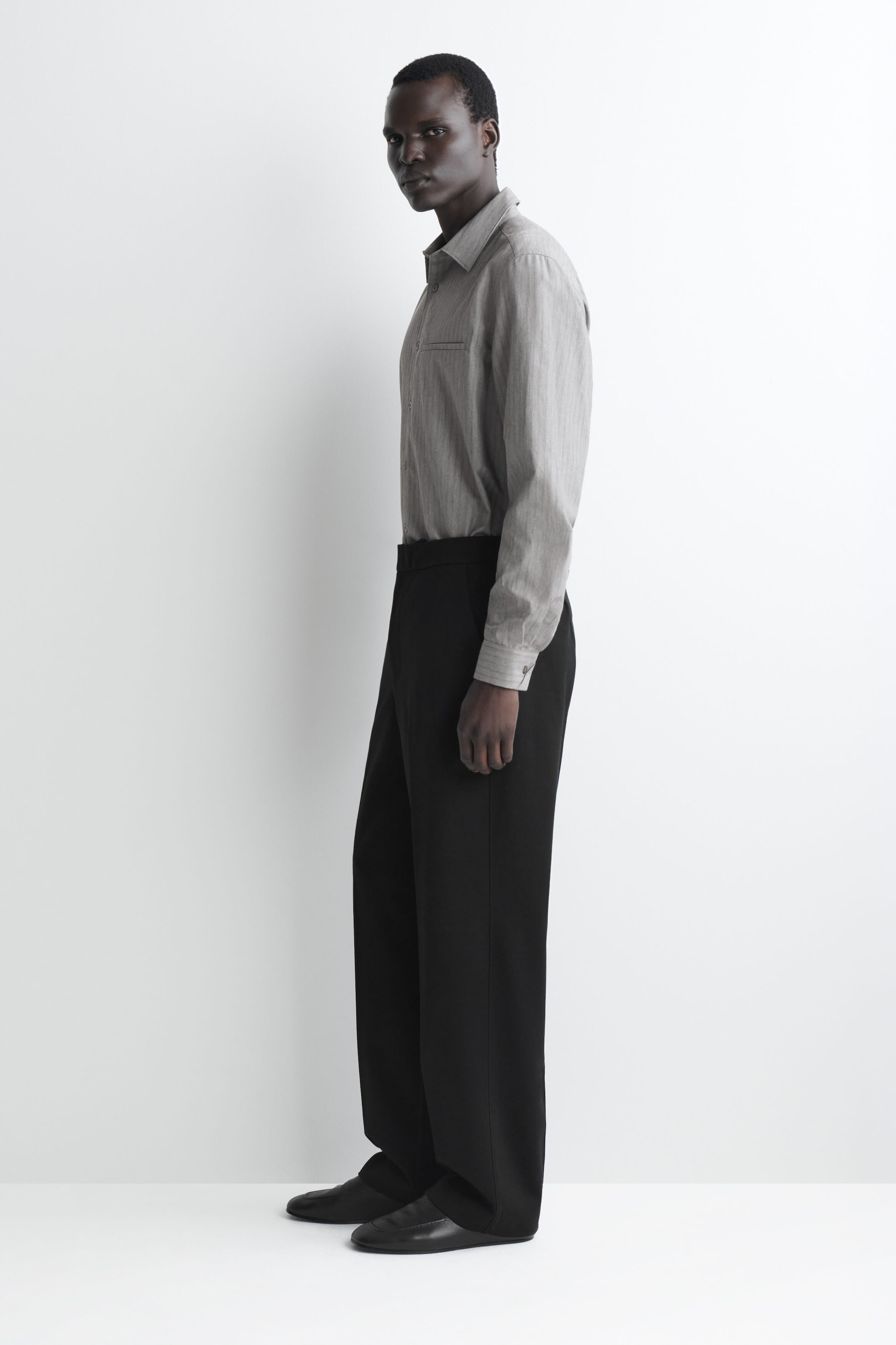 A gray long-sleeve button-up shirt with a point collar and a chest welt pocket is worn tucked into wide-leg black trousers, with the outfit facing left to showcase its relaxed drape. The straight-leg trousers fall over black leather slip-on shoes, highlighting a clean, minimalist silhouette. A gray long-sleeve button-up shirt with a point collar and a chest welt pocket is worn tucked into wide-leg black trousers, with the outfit facing left to showcase its relaxed drape. The straight-leg trousers fall over black leather slip-on shoes, highlighting a clean, minimalist silhouette.