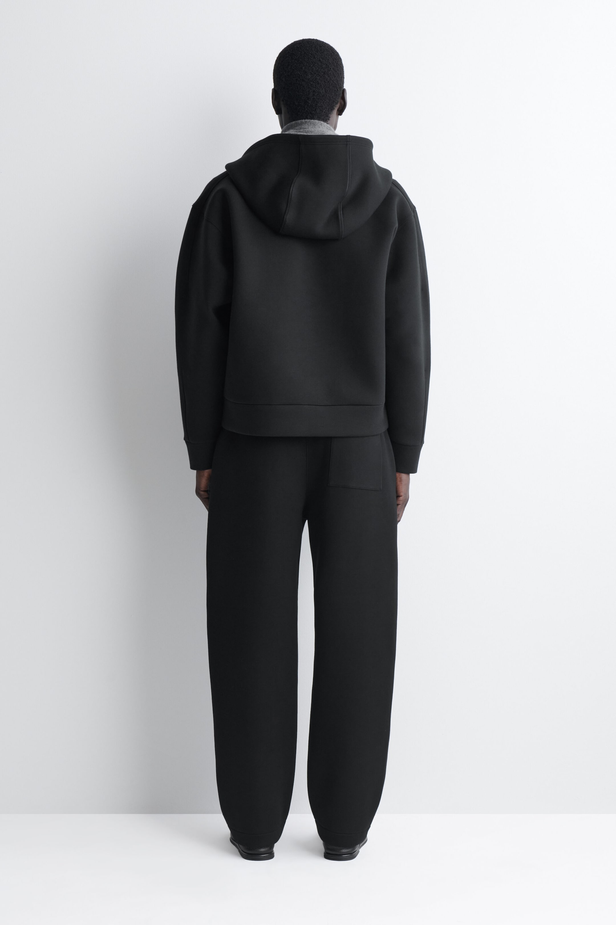 This black matching set is shown from the back, featuring a structured, boxy-fit hoodie with dropped shoulders and a large hood layered over a gray collar. The coordinating wide-leg trousers are styled with a single rear patch pocket, emphasizing the clean, minimalist design of the ensemble.