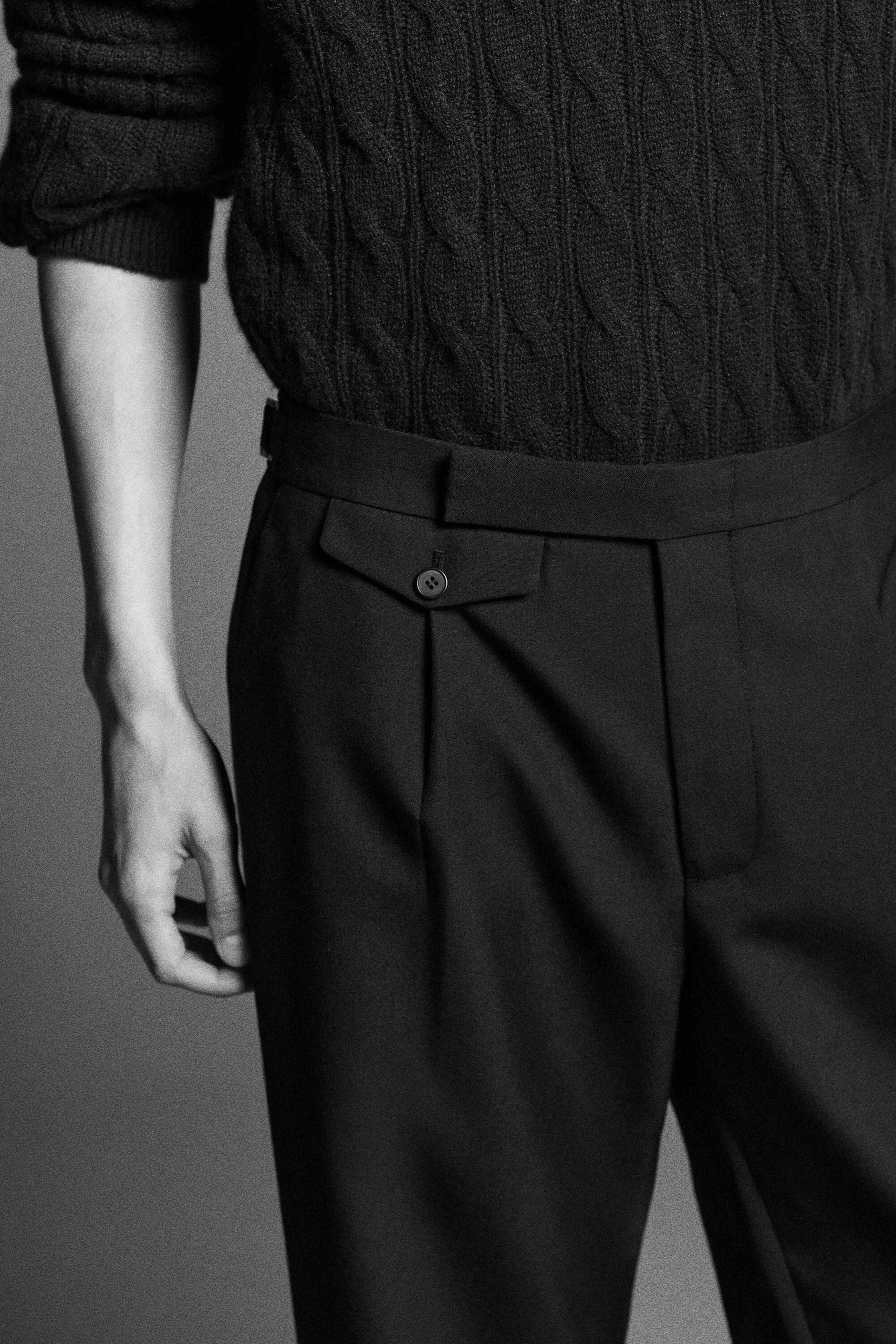 The model is wearing a dark cable-knit cashmere jumper tucked into dark wool slim-fit straight-leg trousers, captured from the front. The jumper features a textured cable-knit pattern with ribbed cuffs, while the trousers showcase a clean waistband with an extended tab and a small buttoned flap pocket.