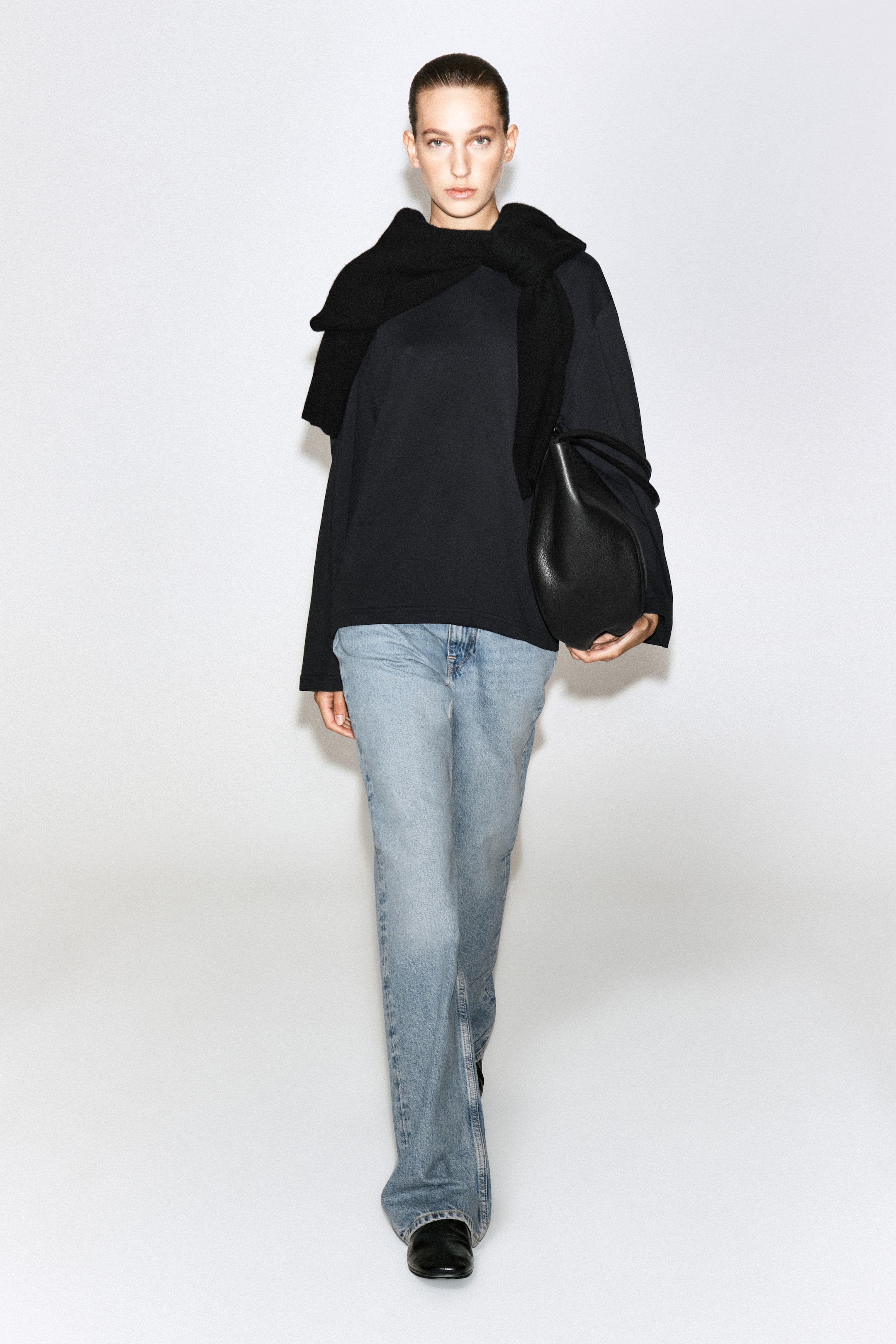 Visualizza immagine più grande: Una maglietta a maniche lunghe in cotone spazzolato nero è indossata dalla modella, abbinata a un maglione nero coordinato appoggiato sulle spalle e a jeans azzurri. La maglietta dalla vestibilità rilassata è abbinata a una borsa in pelle nera portata sotto un braccio.