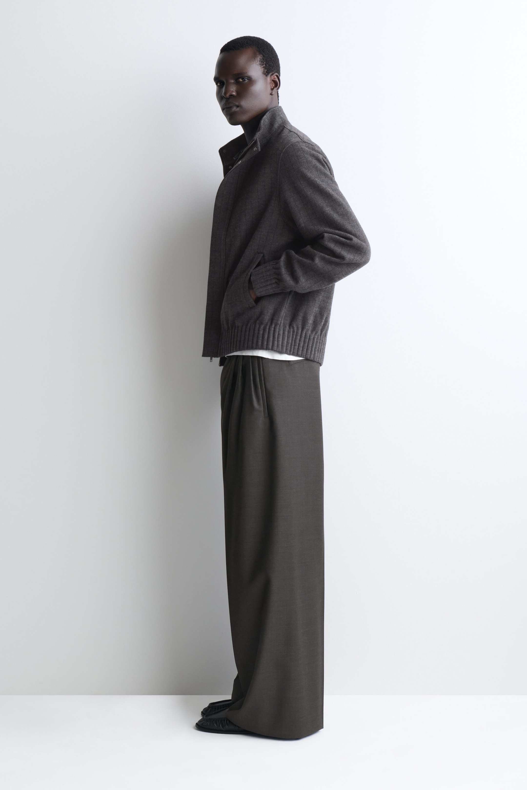 The model, facing left, is wearing a dark gray wool jacket featuring a high funnel neck, ribbed cuffs, and a wide ribbed hem. The jacket is paired with matching dark gray wide-leg trousers that drape to the floor, with a sliver of a white T-shirt peeking out at the hem. The model, facing left, is wearing a dark gray wool jacket featuring a high funnel neck, ribbed cuffs, and a wide ribbed hem. The jacket is paired with matching dark gray wide-leg trousers that drape to the floor, with a sliver of a white T-shirt peeking out at the hem.