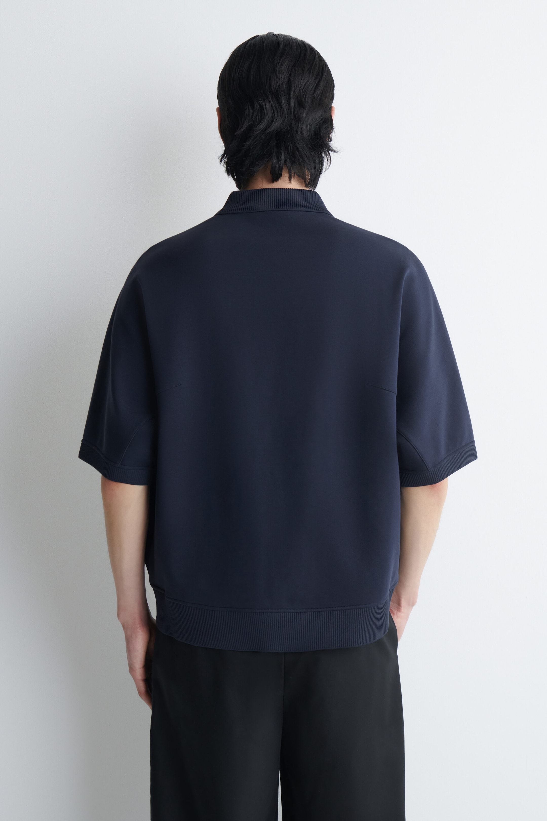The model is wearing a navy blue scuba jersey polo shirt, shown from behind to highlight its structured design. The short-sleeved shirt features a ribbed collar, ribbed sleeve cuffs, and a wide ribbed hem. The model is wearing a navy blue scuba jersey polo shirt, shown from behind to highlight its structured design. The short-sleeved shirt features a ribbed collar, ribbed sleeve cuffs, and a wide ribbed hem.