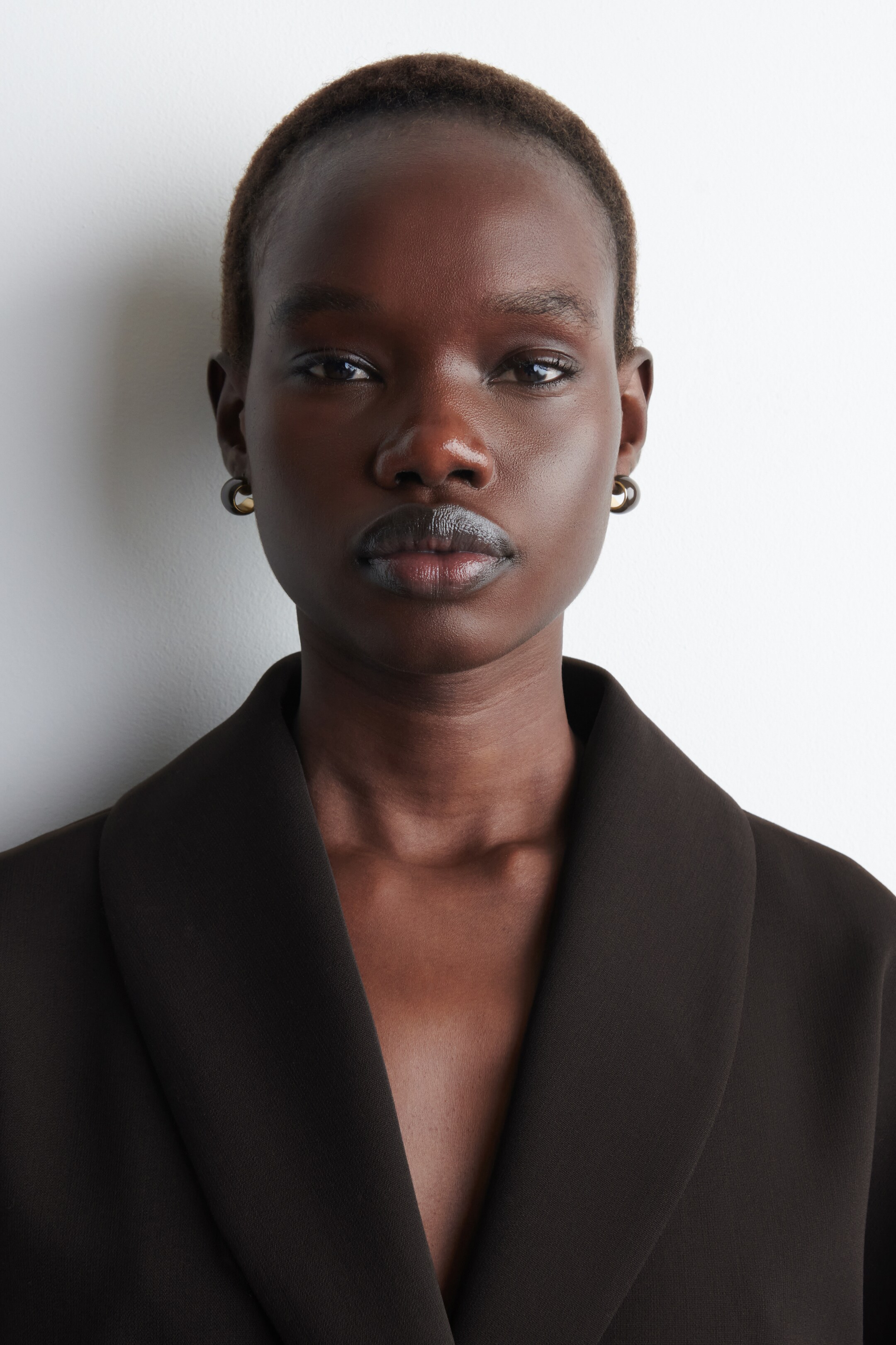 Größeres Bild ansehen: Ein dunkelbrauner Blazer präsentiert breite Schalkragen und eine offene Vorderseite. Kleine, zweifarbige Creolen sind sichtbar, mit einem dunklen Außenring und einem goldenen Halbmond-Detail.