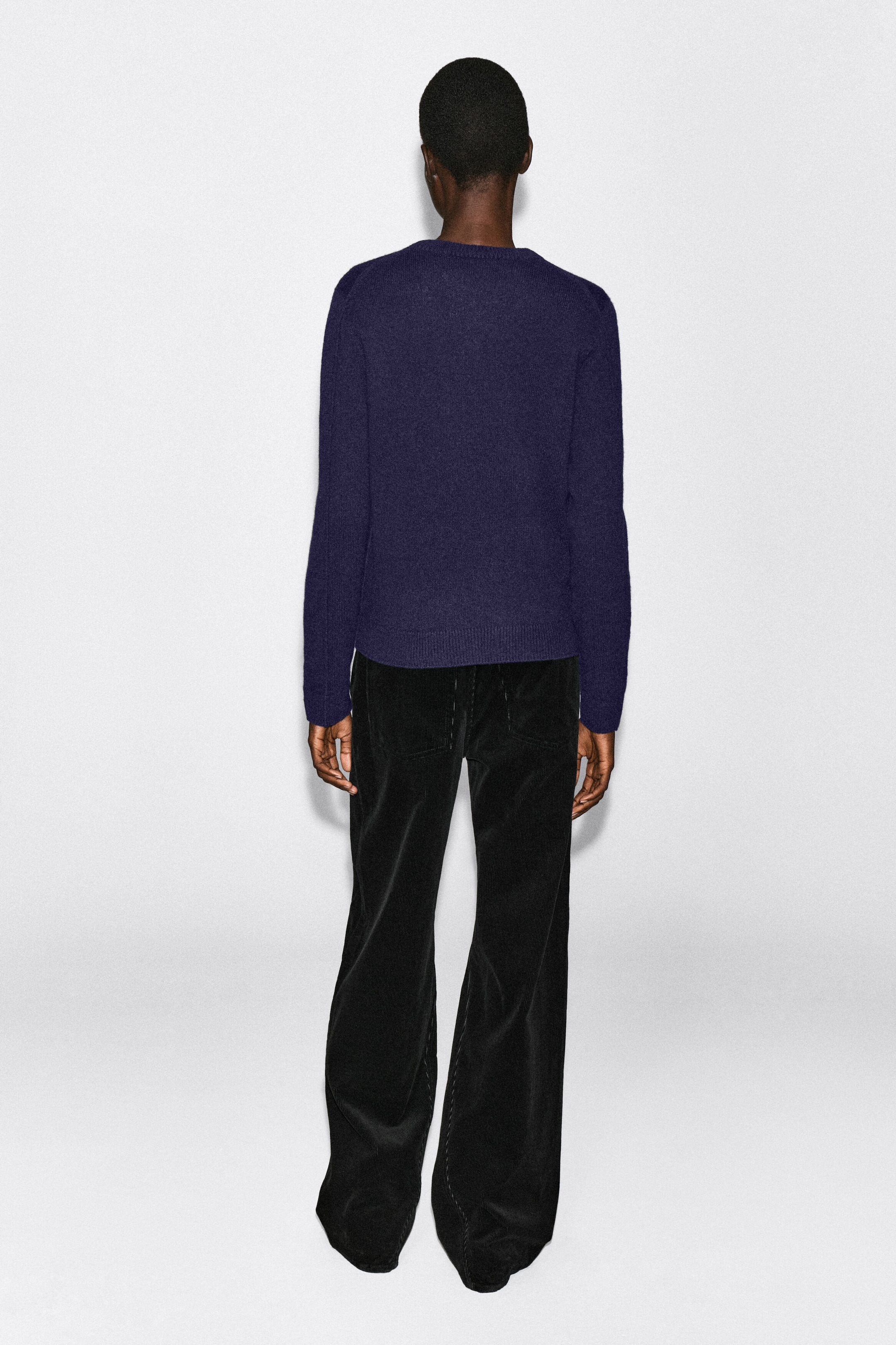 Un pull en cachemire bleu foncé est porté par le mannequin, vu de dos. Ce vêtement en maille à manches longues présente un ourlet côtelé et est associé à un pantalon noir à jambes larges. Un pull en cachemire bleu foncé est porté par le mannequin, vu de dos. Ce vêtement en maille à manches longues présente un ourlet côtelé et est associé à un pantalon noir à jambes larges.