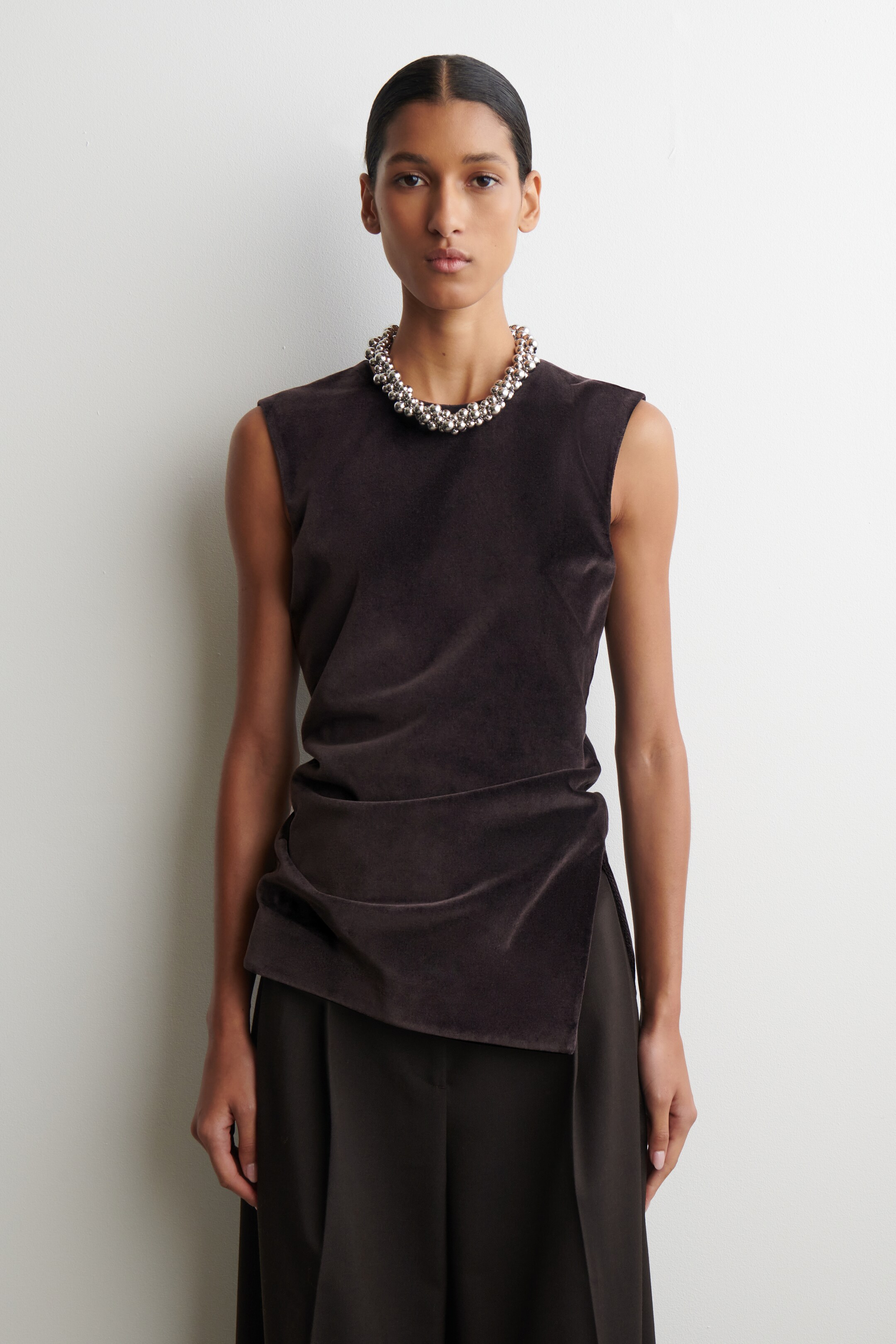 Grösseres Bild ansehen: Ein dunkelbraunes ärmelloses Top ist nach vorne gerichtet und verfügt über einen runden Ausschnitt sowie ein asymmetrisches Drapierungsdesign, bei dem der Saum sich um die Taille wickelt. Es wird über dunkelbraunen Hosen gestylt, und eine silberfarbene, perlenbesetzte Halskette schmückt den Ausschnitt.