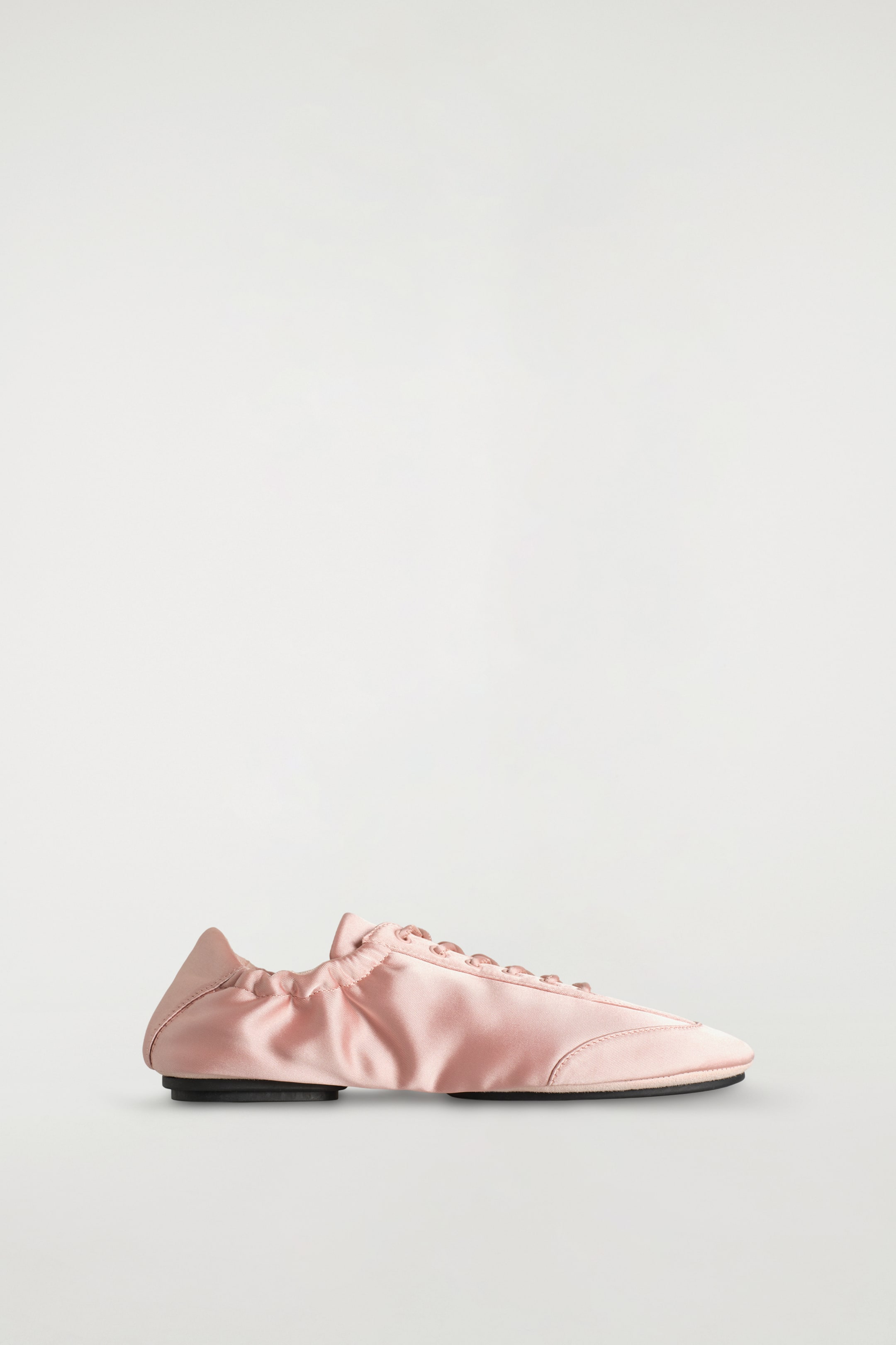 Agrandir l'image: Une basket de ballet rose clair est présentée de profil, avec une tige en satin à l'éclat doux, agrémentée de touches de daim sur le bout et le long du laçage. L'arrière de la chaussure présente un détail froncé et élastiqué, reposant sur une semelle plate noire.