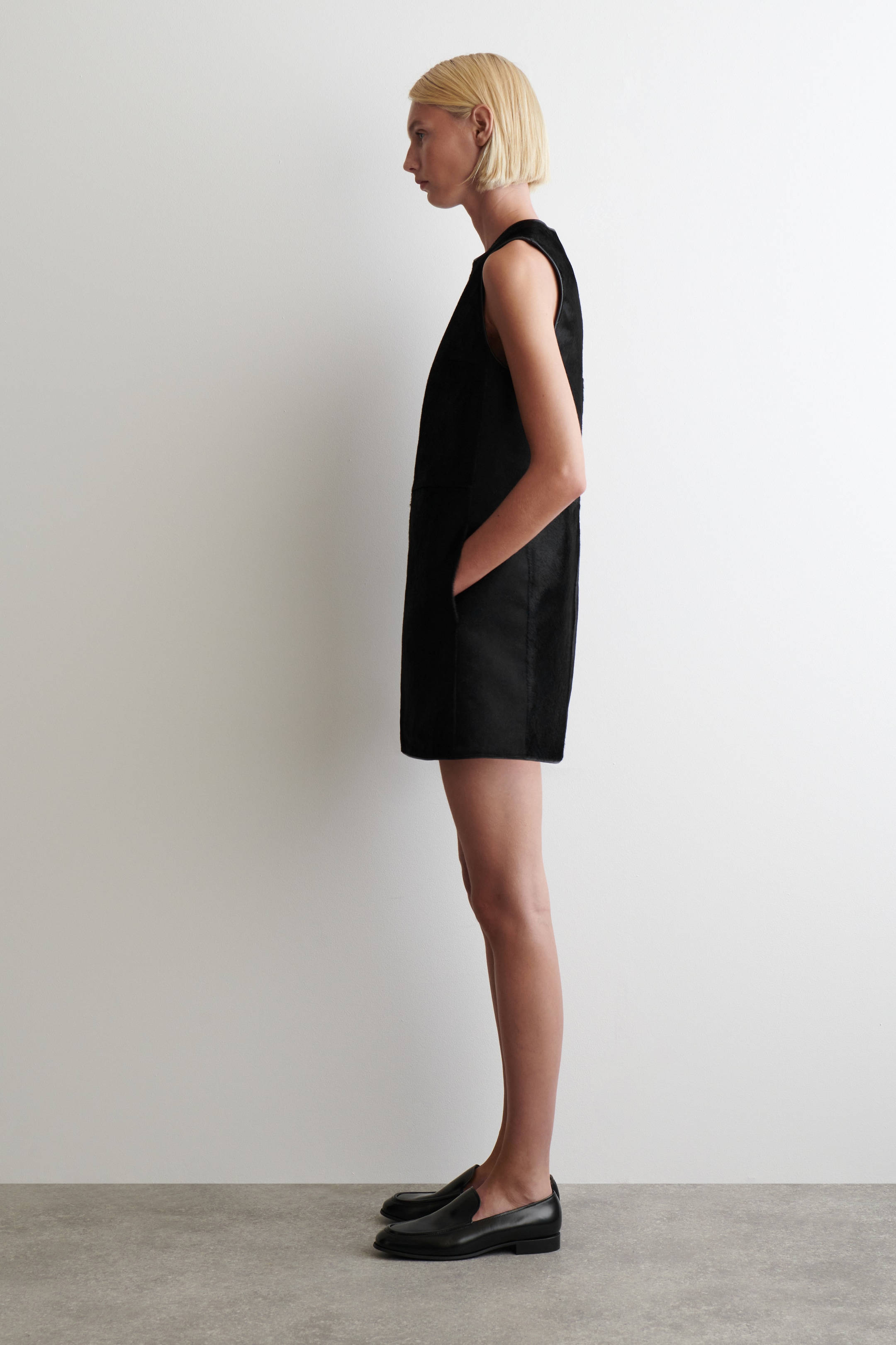Agrandir l'image: Une robe droite sans manches noire est présentée de son profil gauche. Elle présente un col haut et des poches latérales visibles, avec une main reposant à l'intérieur d'une poche, et est coupée au-dessus du genou.