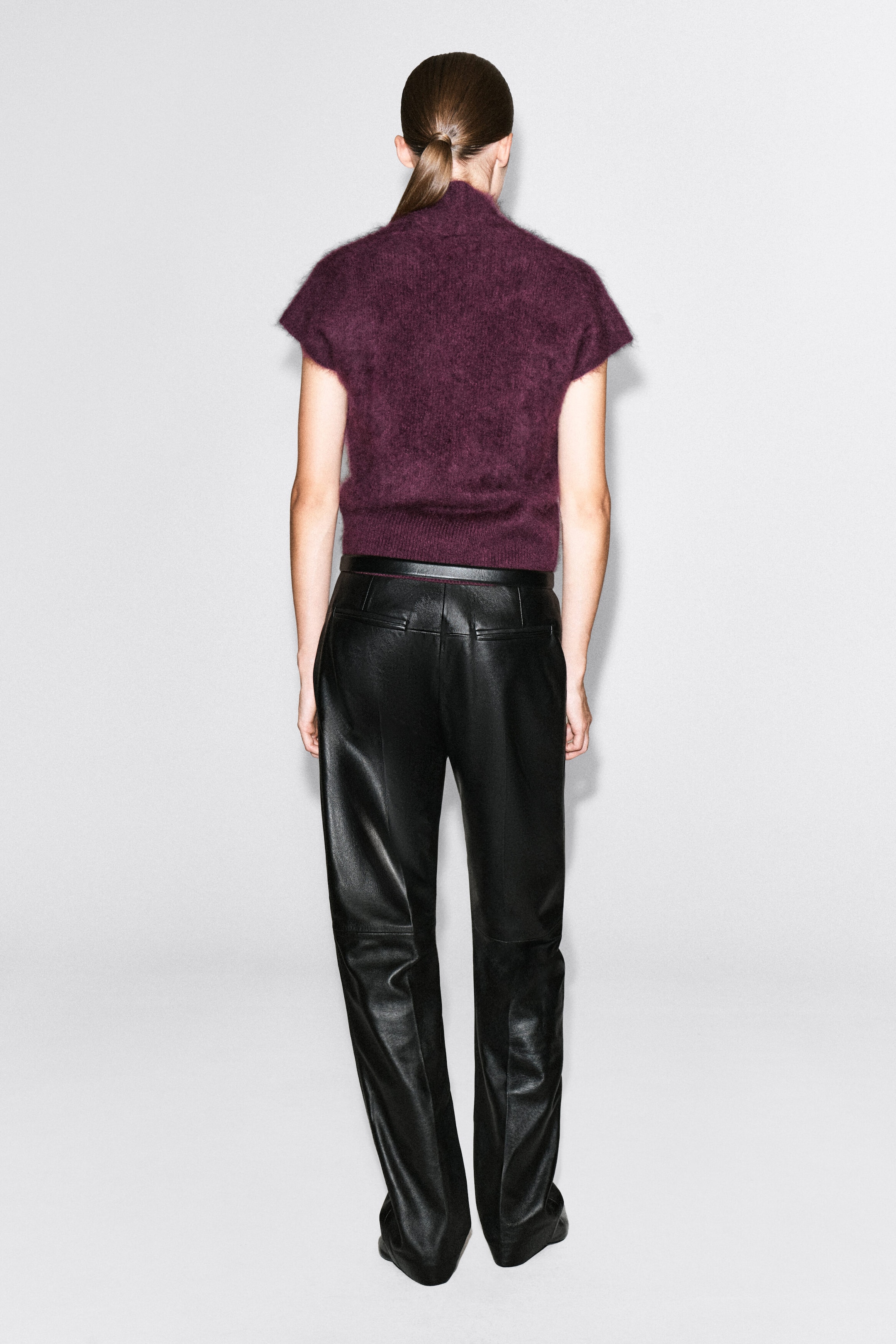 A draped brushed merino wool top in deep purple is worn by the model, shown from the back. The fuzzy, textured knit top features cap sleeves and a ribbed hem, and is styled with black leather trousers.