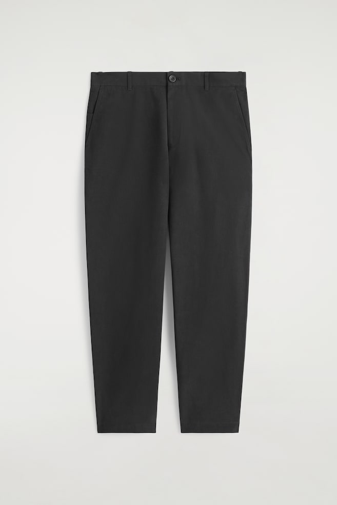 Ein Paar schwarze Chinos wird flach von vorne gezeigt, mit einem geraden Bein. Sie zeigen einen Bund mit Gürtelschlaufen, einen einzelnen Knopfverschluss und schräge Taschen an den Seiten. - 1