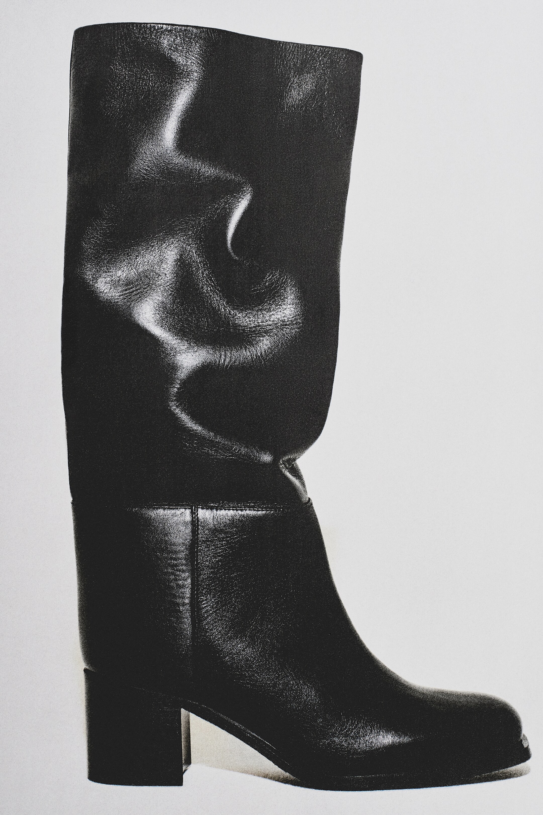 Größeres Bild anzeigen: Ein schwarzer Lederstiefel in Kniehöhe wird von der Seite gezeigt, mit einer mandelförmigen Spitze, einem breiten Schaft, der natürliche Falten aufweist, und einem Blockabsatz.