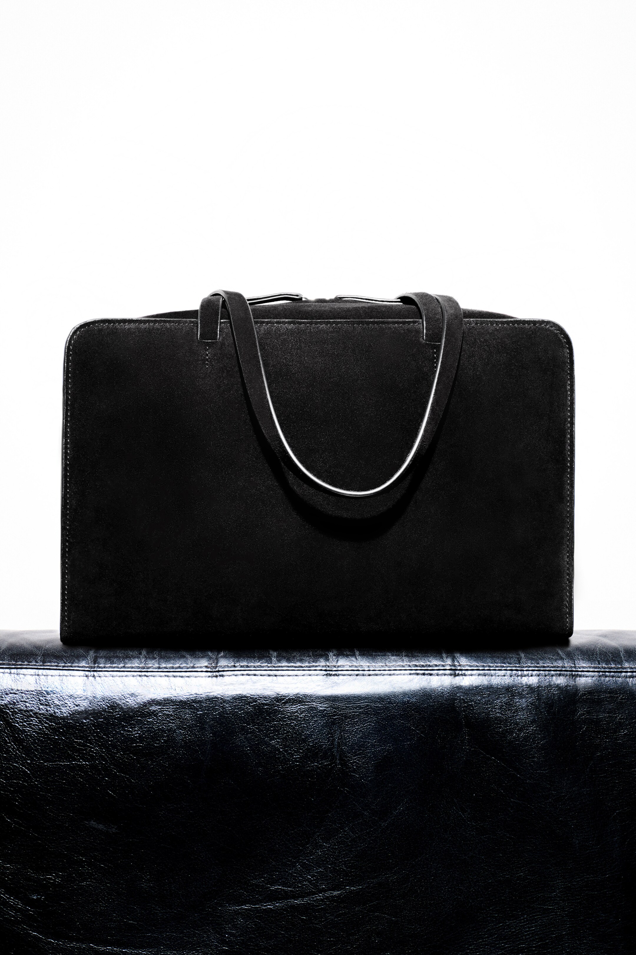 Größeres Bild anzeigen: Eine dunkle Wildleder-Bowlingtasche wird von vorne präsentiert und zeigt eine kastige Silhouette. Sie verfügt über verlängerte flache Träger mit hellen Kanten und einen Reißverschluss entlang der Oberseite.