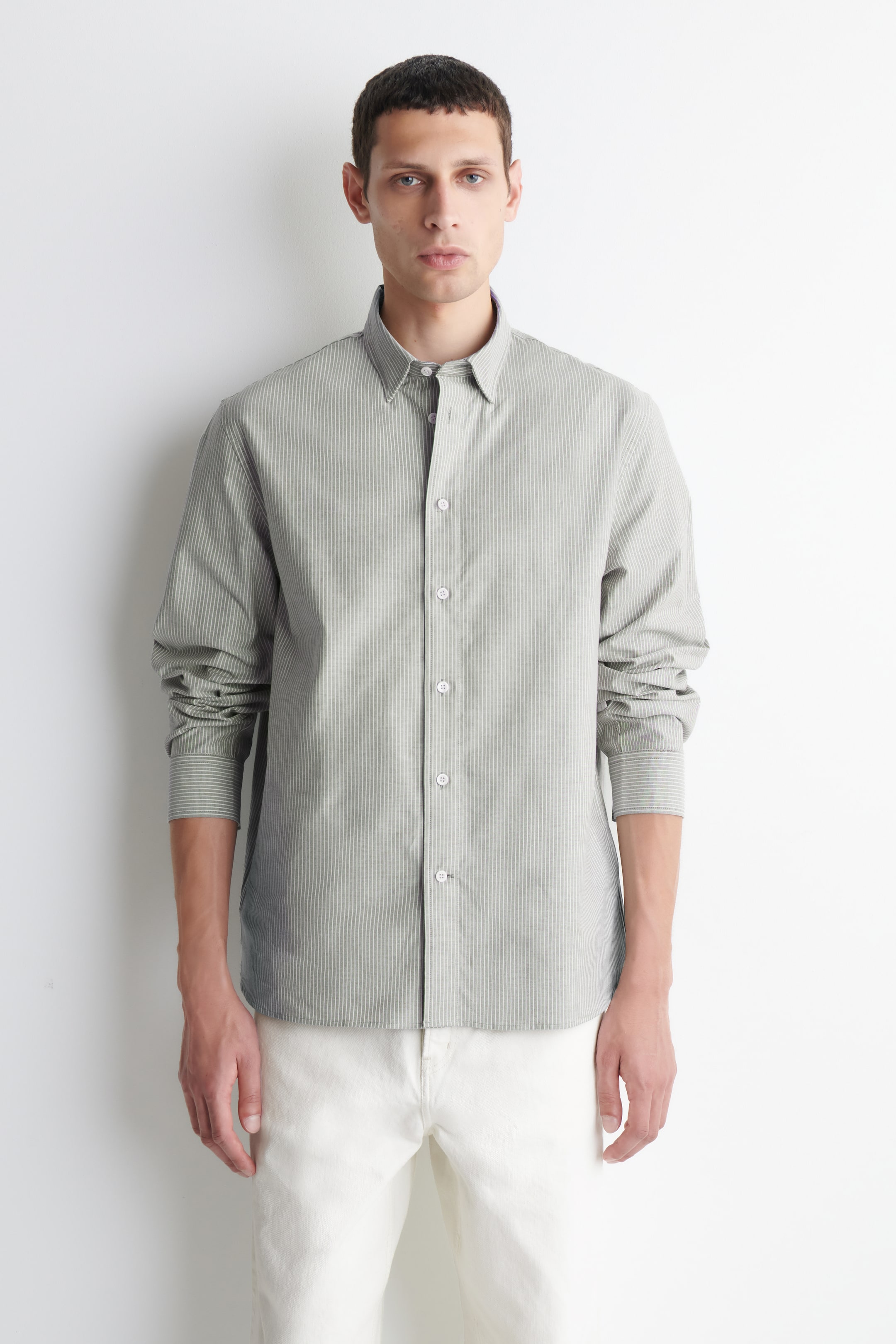 View larger image: A muted green-grey button-up shirt is displayed facing forward, featuring fine vertical stripes, a button-down collar, and a full button placket with white buttons. Its long sleeves are pushed up, creating folds above the cuffs.
