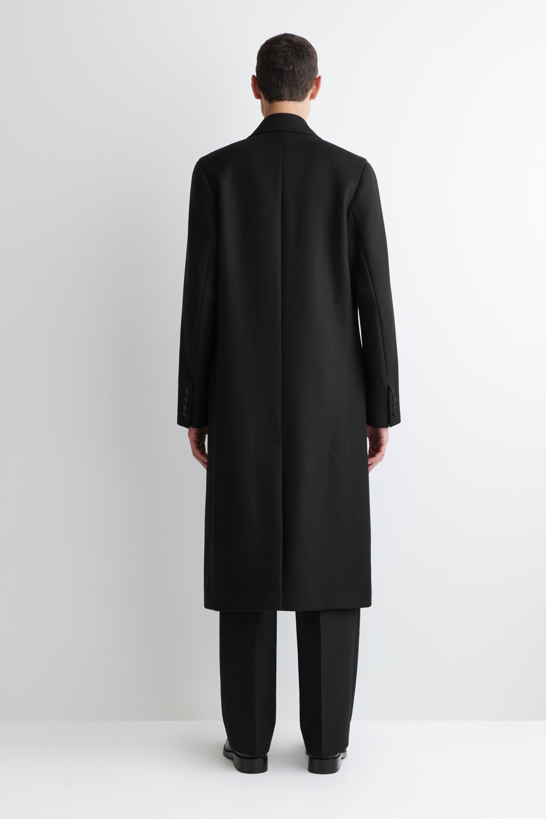 Visualizza immagine più grande: Un lungo cappotto nero viene visto dal retro, mostrando la sua lunghezza completa e la silhouette dritta. Presenta un colletto classico, maniche lunghe con quattro bottoni su ciascun polsino e uno spacco singolo al centro della schiena, che si estende fino a metà polpaccio.