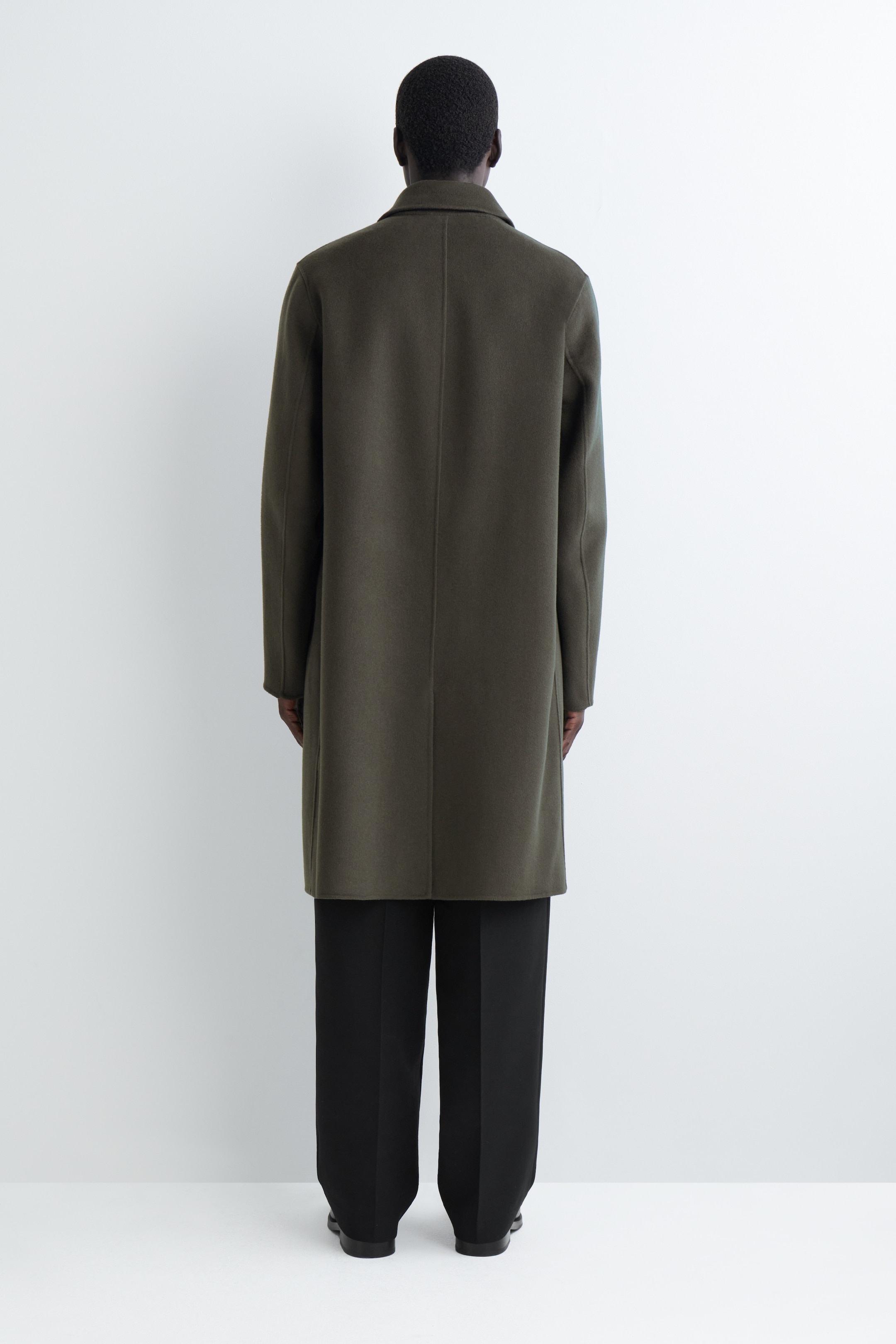 Visualizza immagine più grande: Un cappotto verde oliva è rivolto lontano dall'osservatore, mostrando il suo retro. Il cappotto presenta un colletto strutturato e una cucitura centrale sul retro, e ha una vestibilità dritta che si estende fino a poco sopra il ginocchio.